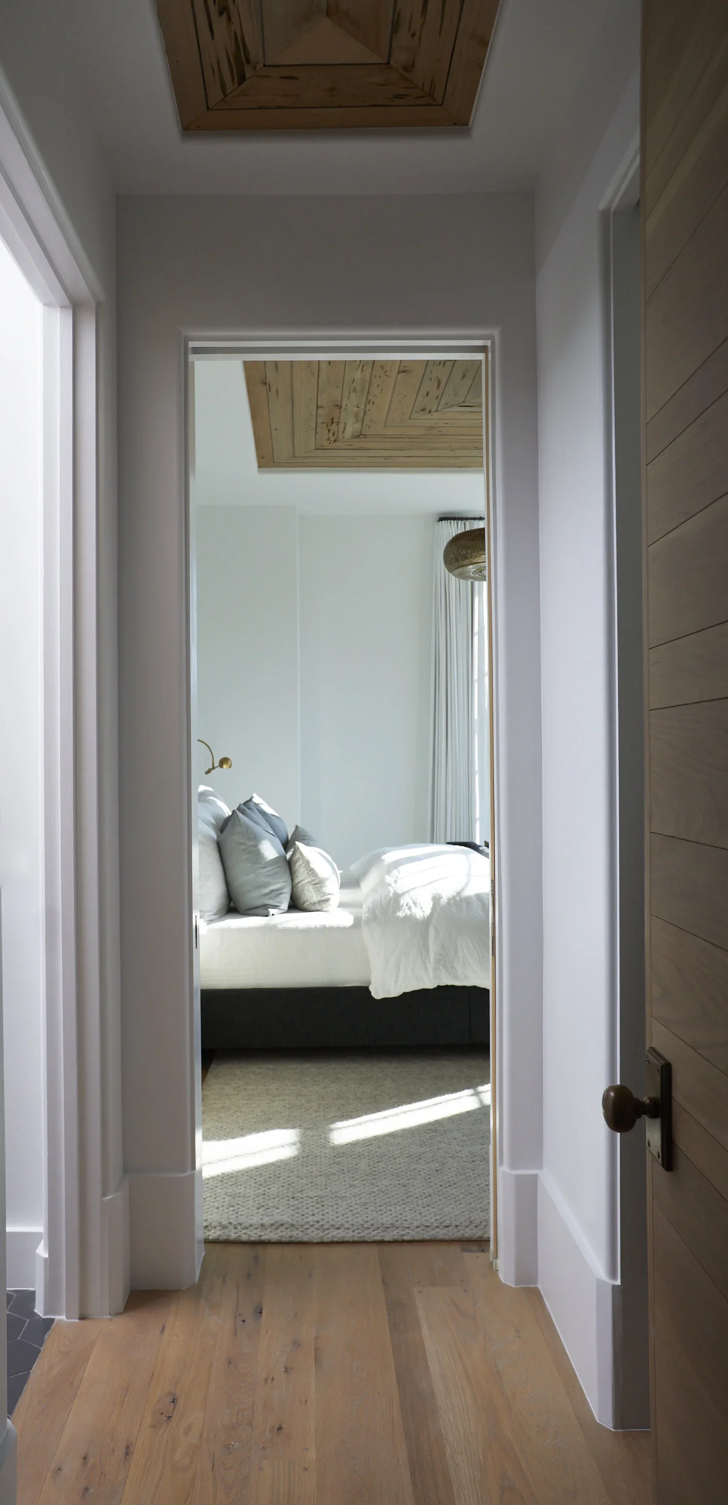 A view of a bedroom through an open doorway, with a bed featuring white bedding and multiple pillows, light coming through a window with curtains, and a wooden ceiling.