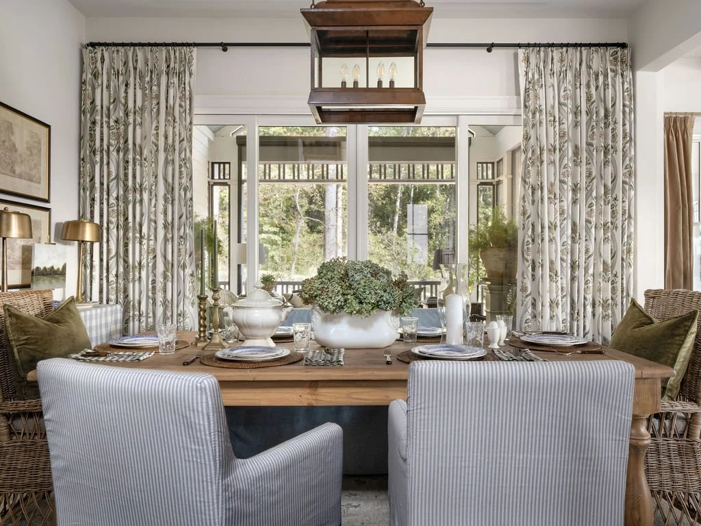 A dining room with a large wooden table set for a meal, surrounded by various chairs including upholstered armchairs and wicker chairs. The table has a centerpiece with greenery, white vases, and dishes. Behind the table are large windows with sheer,