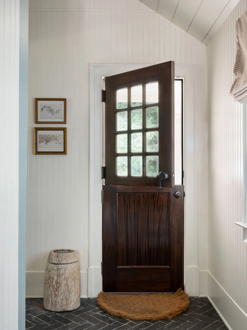 Interior of a house entryway with a dark wooden door with glass panes, framed by white walls with vertical paneling, a small circular stone stool on a dark tiled floor, two framed botanical prints on the wall, and a small window with a beige curtain.
