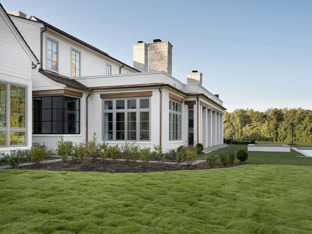 White residential house with large windows, multiple chimneys, and a well-maintained lawn in a rural setting with trees in the background.