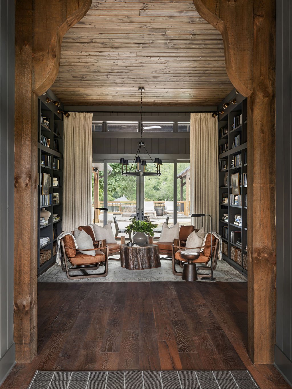 Cozy living room with leather armchairs, a rustic coffee table, and tall bookshelves, overlooking a backyard through large windows.