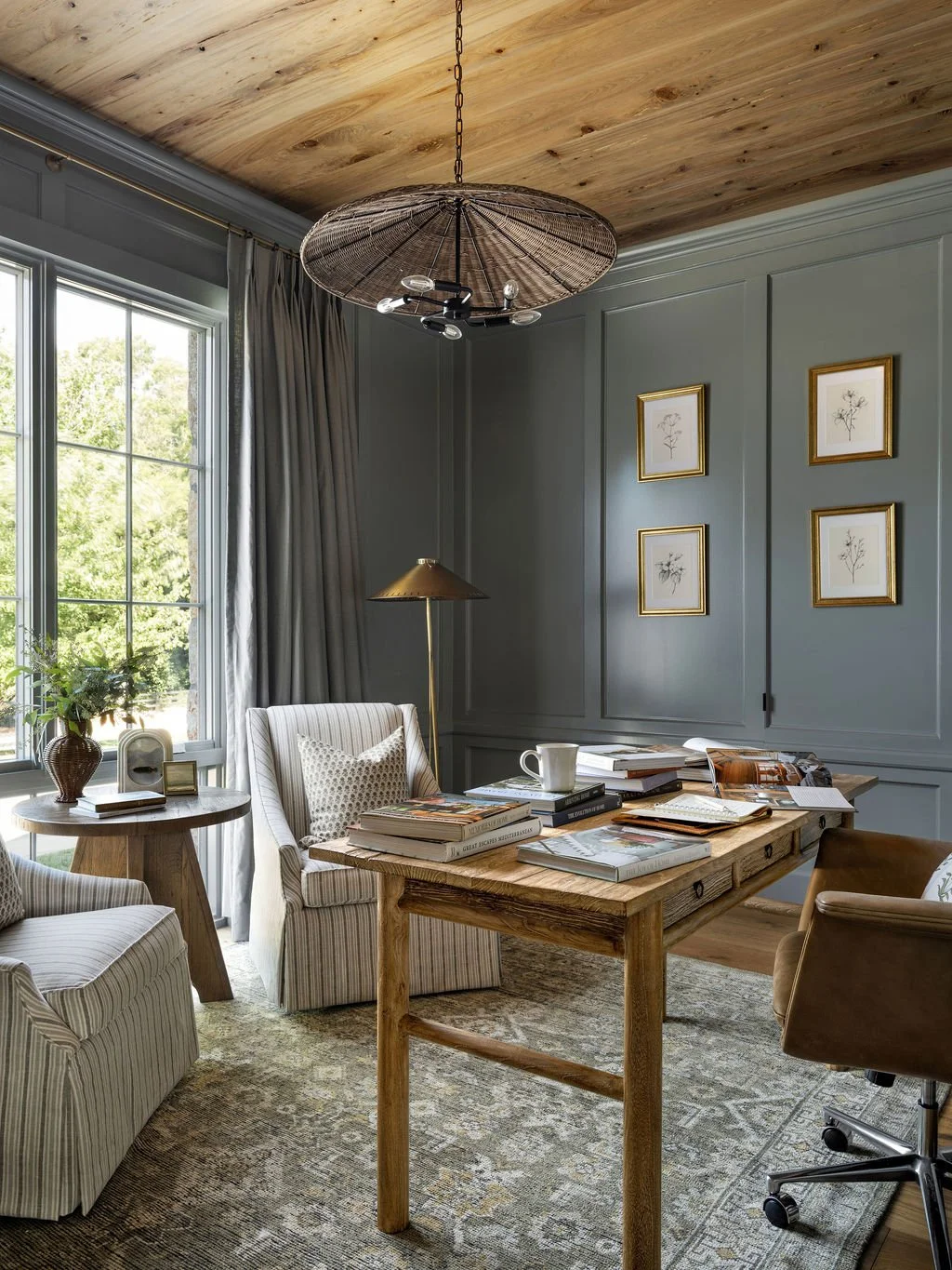 A cozy home office with a wooden desk topped with open books and a coffee mug, a striped armchair with a decorative pillow, and a side table with a potted plant and a clock, illuminated by natural light from a large window with gray curtains. The roo