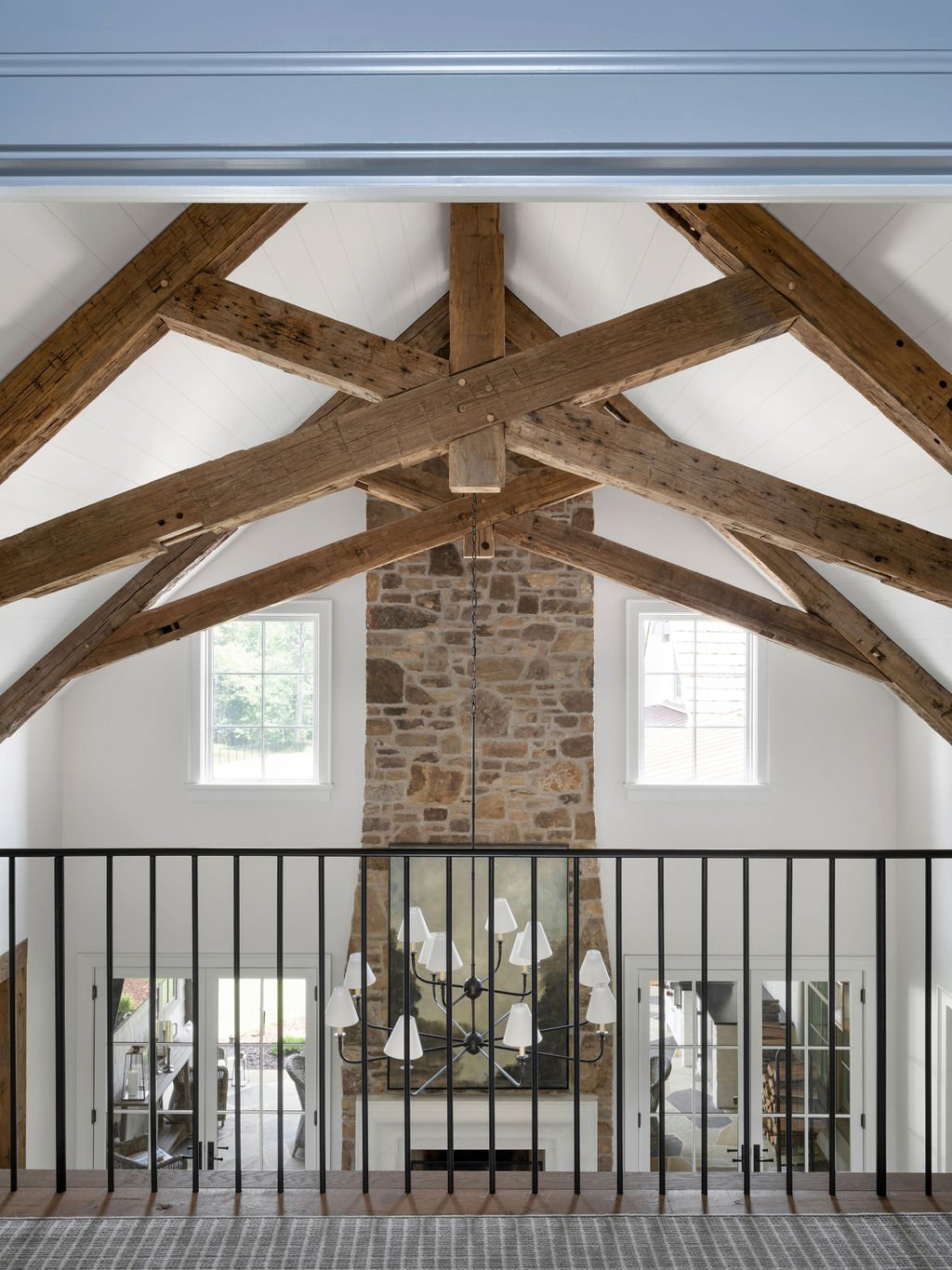 Interior view of a loft with exposed wooden beams, a brick fireplace, two large windows, a chandelier, and a view into a living room area.