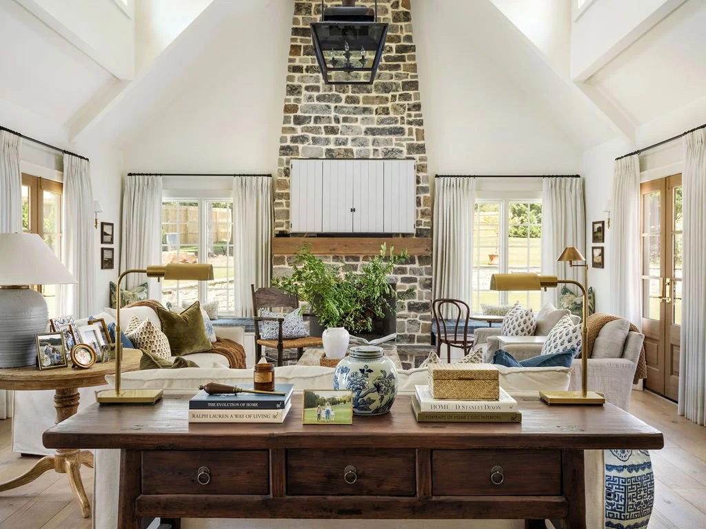 Living room with white walls, high vaulted ceiling, stone fireplace, and large windows with white curtains, furnished with sofas, armchairs, and decorative pillows.