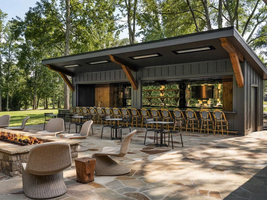 An outdoor bar and seating area surrounded by trees, with a modern black building featuring a row of barstools, tables, and chairs on a stone patio, and a fire pit with chairs nearby.