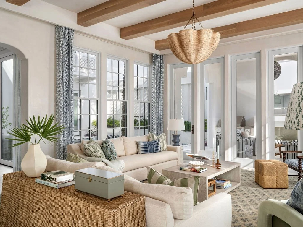 Bright living room with beige sofa, patterned pillows, wicker side table with plants, coffee table with books and decor, large windows with curtains, and a hanging light fixture.