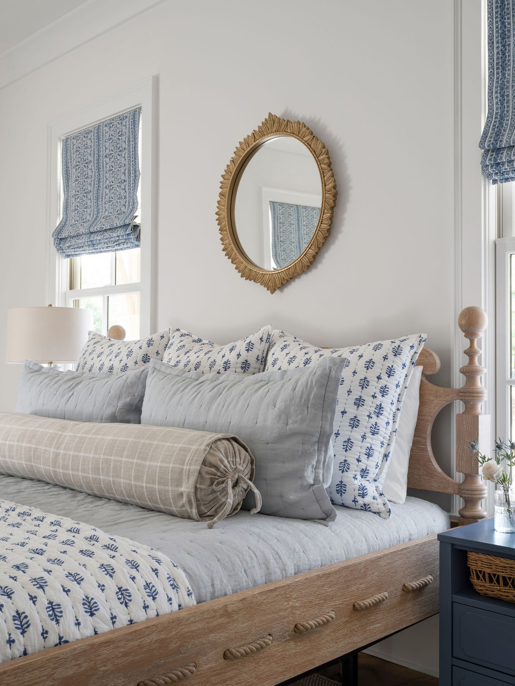 A cozy bedroom with a wooden bed, featuring a beige and white striped bolster pillow and blue and white patterned pillows. A round gold-framed mirror hangs on the white wall above the bed. There are windows with blue patterned Roman shades and a nigh
