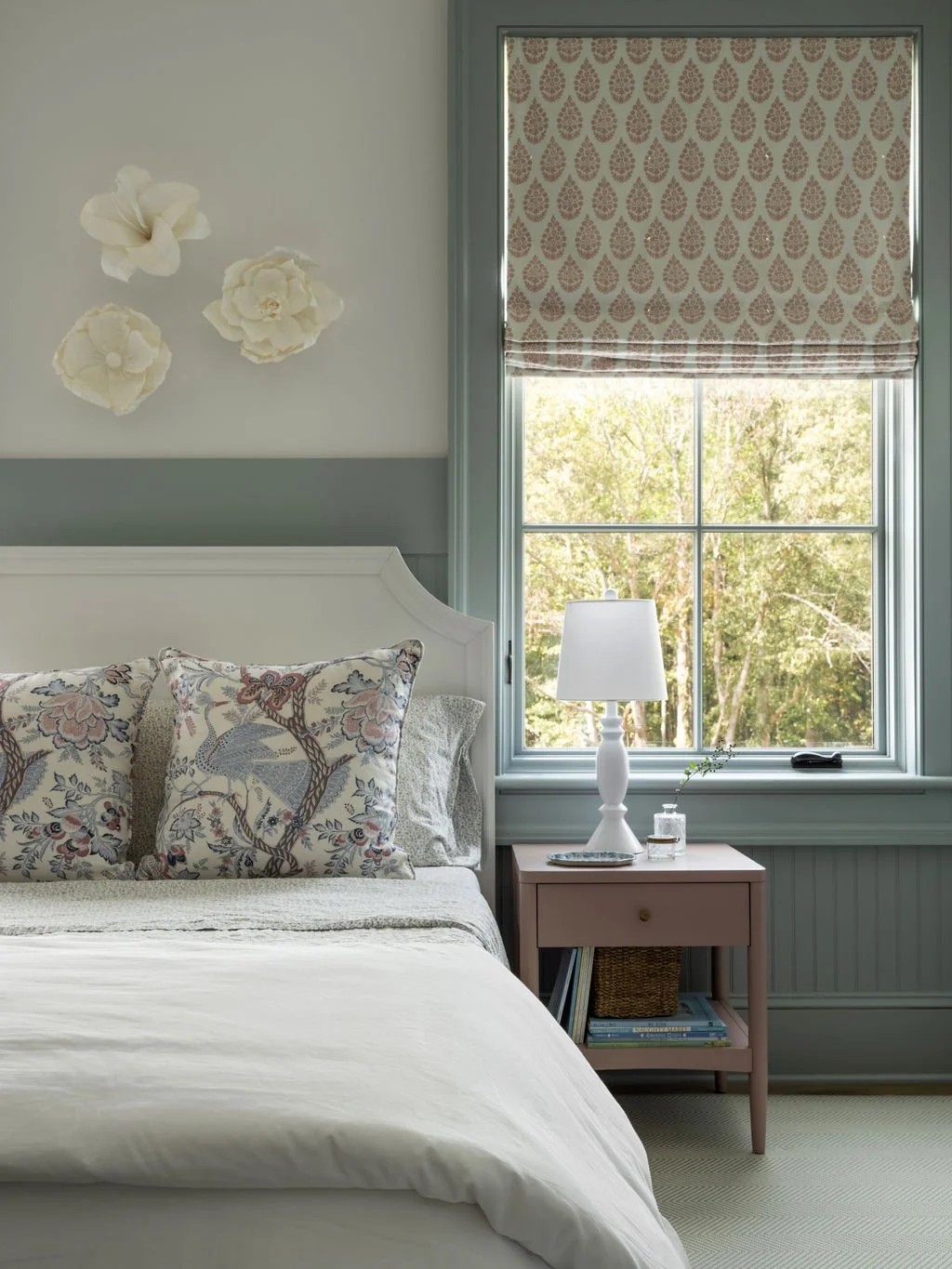 Bedroom with a white bed, floral pillows, pink nightstand, table lamp, window with patterned Roman shade, and decorative wall flowers.
