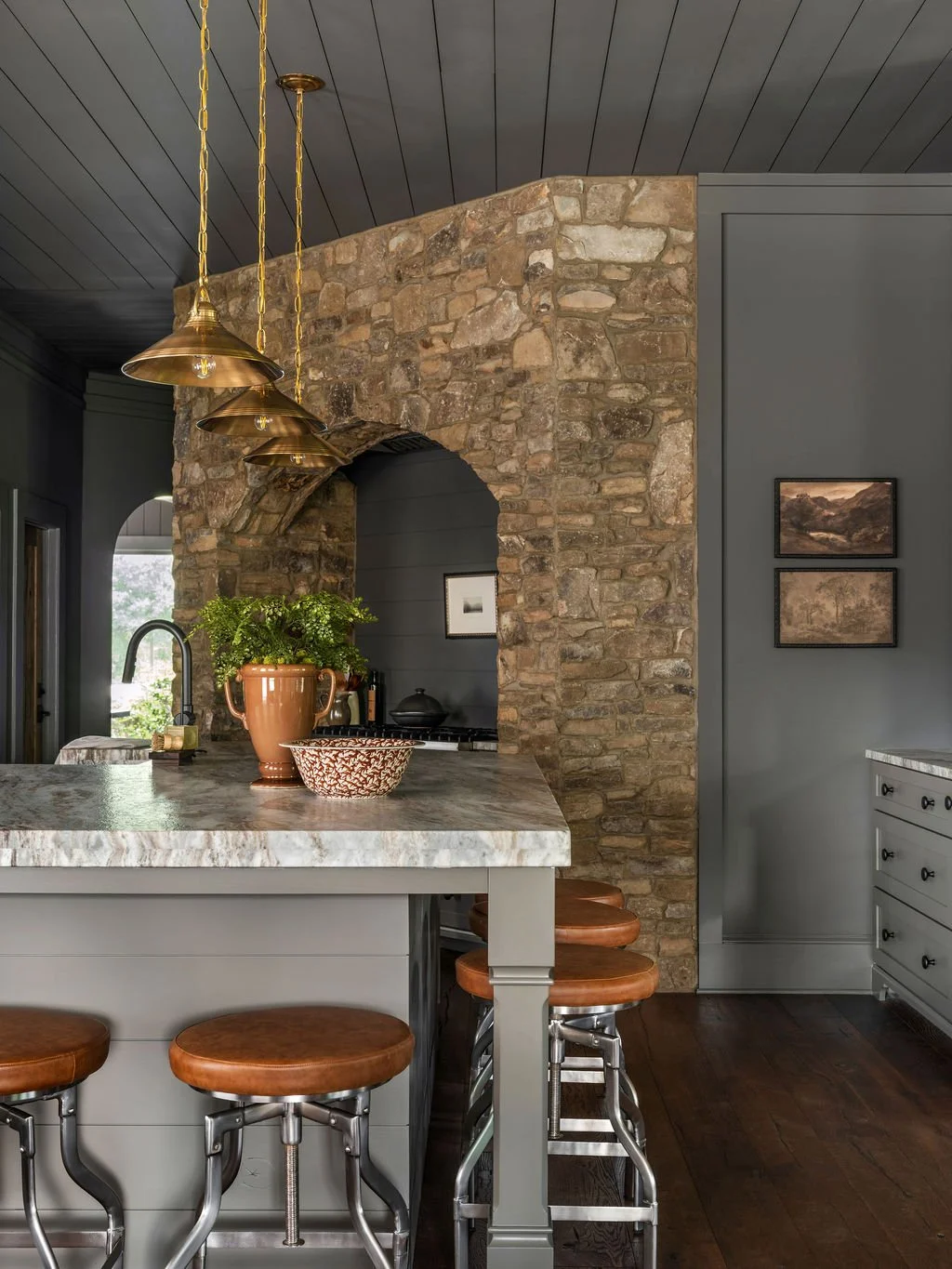 Contemporary kitchen with a stone and brick wall, gray cabinetry, marble countertop, three leather stools, brass pendant lights, and a window with greenery outside.