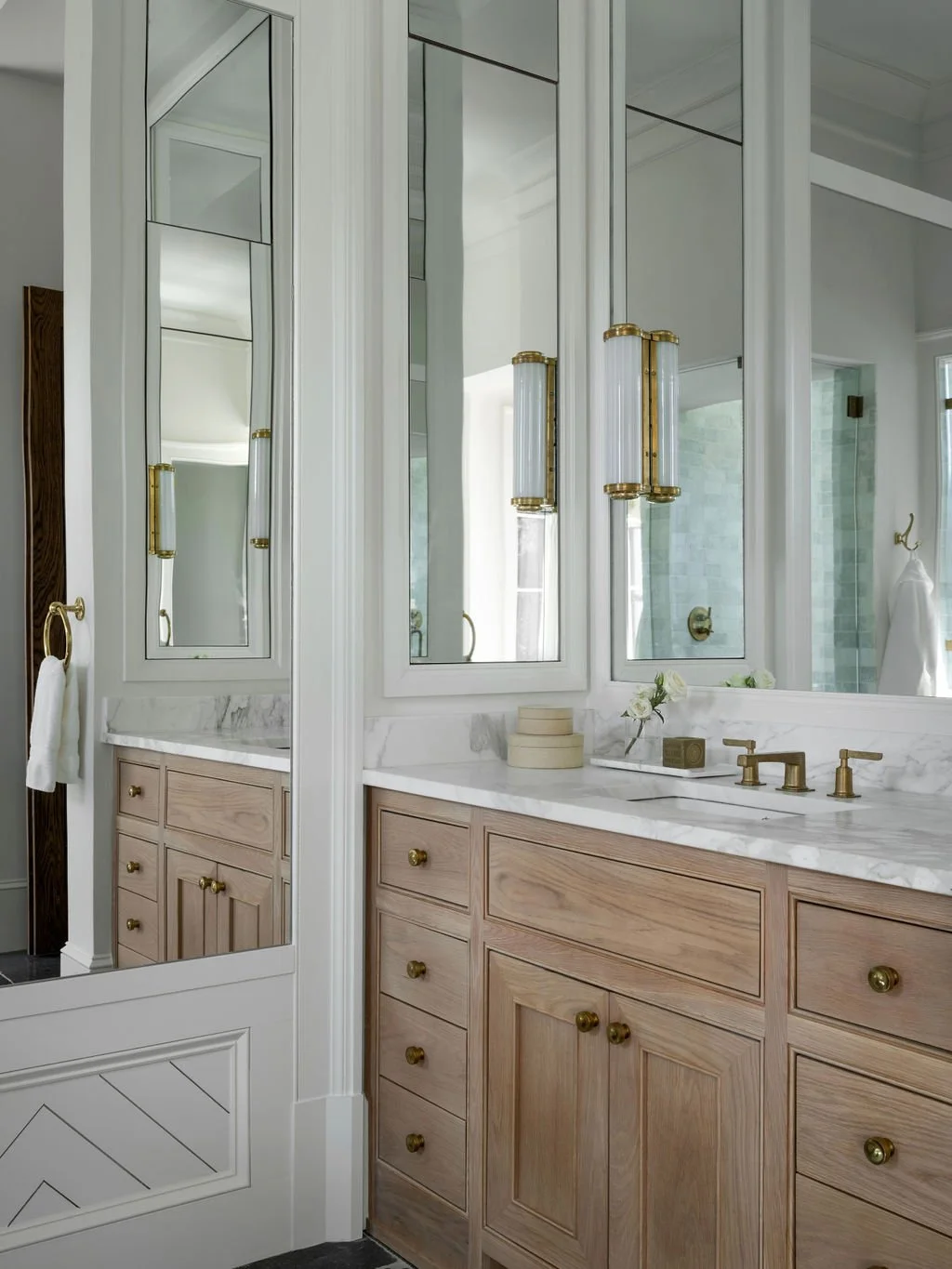 A bathroom vanity with a marble countertop, wooden cabinets with brass knobs, and large mirrored medicine cabinets. There are brass fixtures at the sink and decorative light fixtures on either side of the mirror.