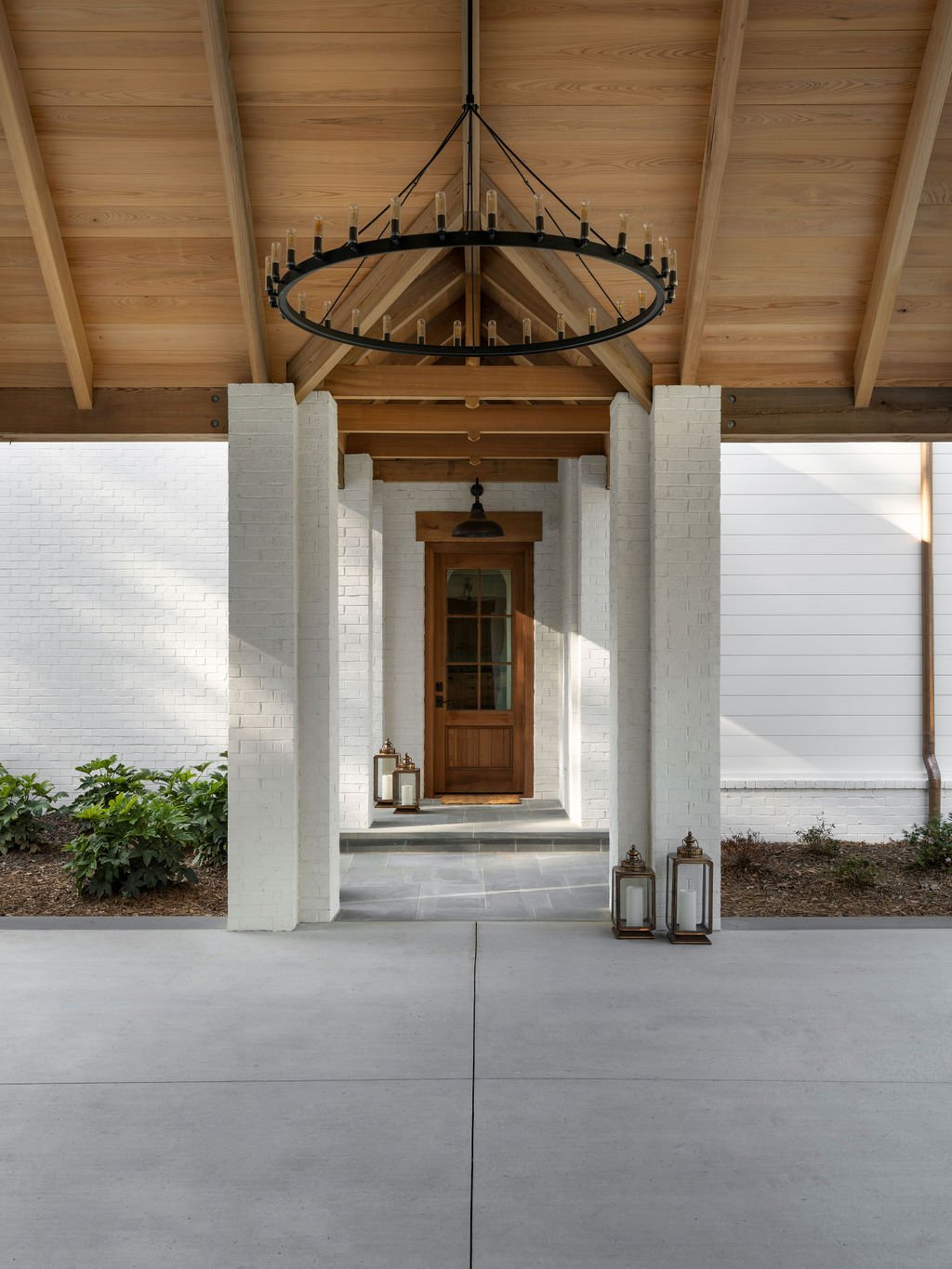 Covered porch with white brick pillars, wooden roof, black chandelier, potted lanterns, and a wooden front door, with some plants on either side.