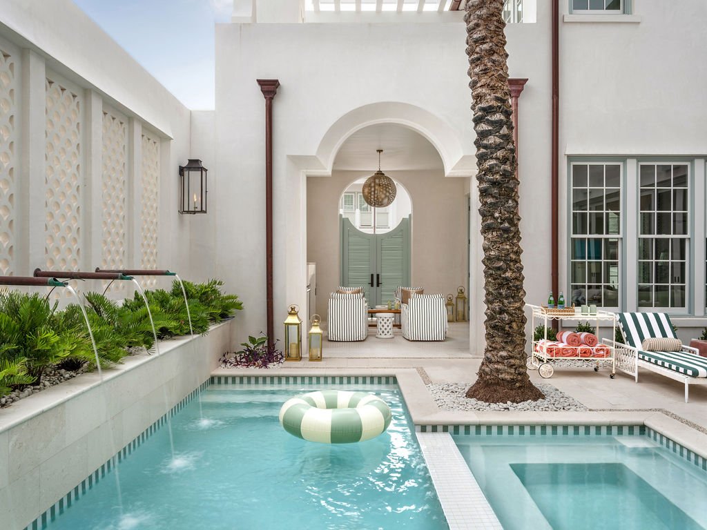 A luxurious outdoor pool area with a small hot tub, a white house with large windows, striped chairs, pool floats, potted plants, and a palm tree.