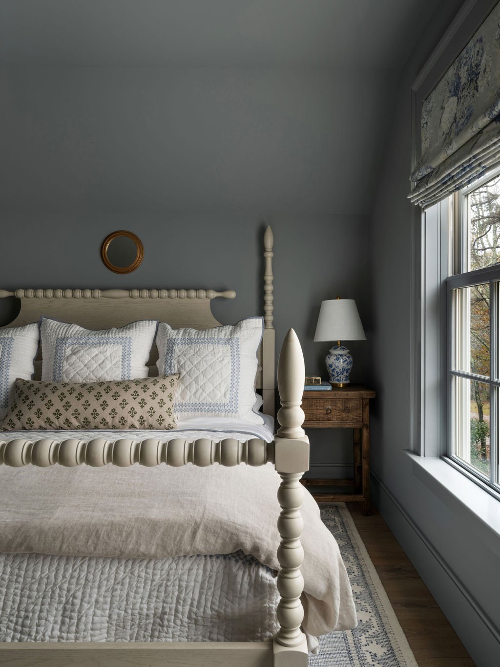 A bedroom with a cream-colored four-poster bed, white quilted pillows, a wooden nightstand with a blue and white ceramic lamp, a window with patterned curtains, and a small round mirror on the gray wall.