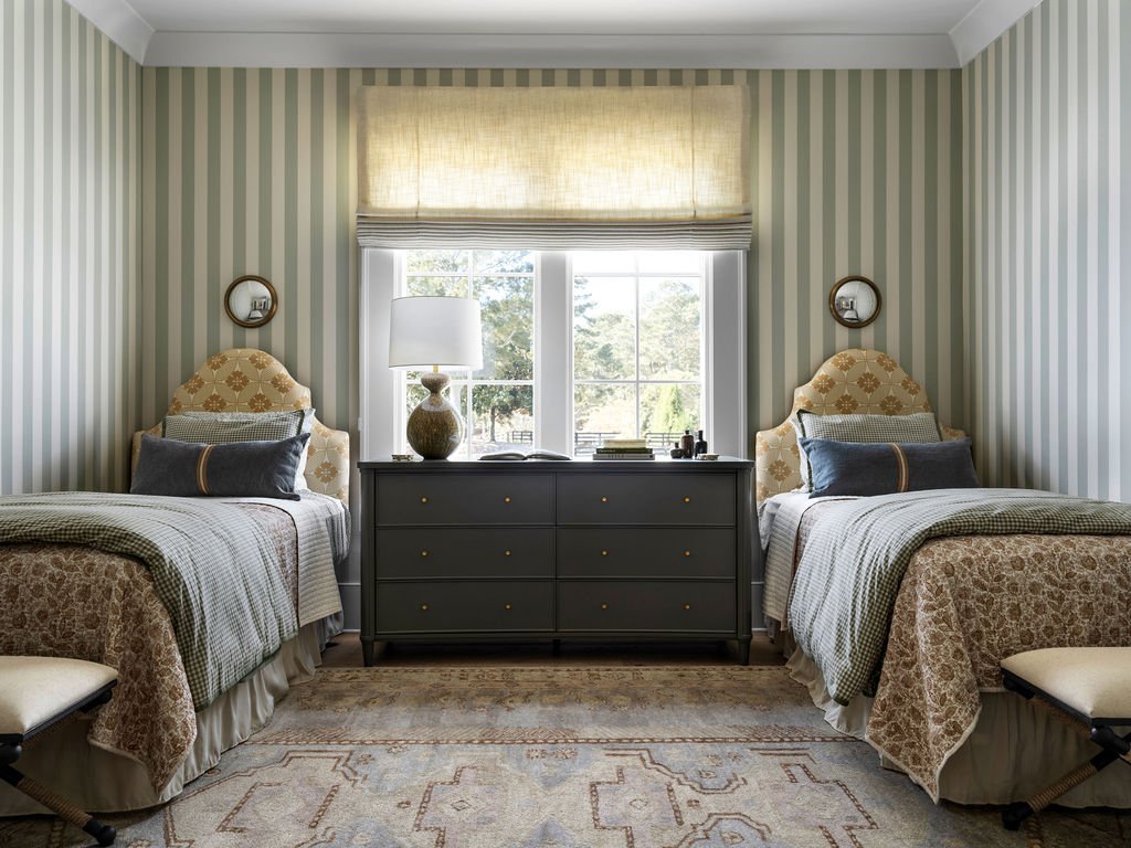 A bedroom with two twin beds, patterned headboards, striped walls, window with a yellow shade, and a dark dresser between the beds decorated with a lamp, books, and bottles.