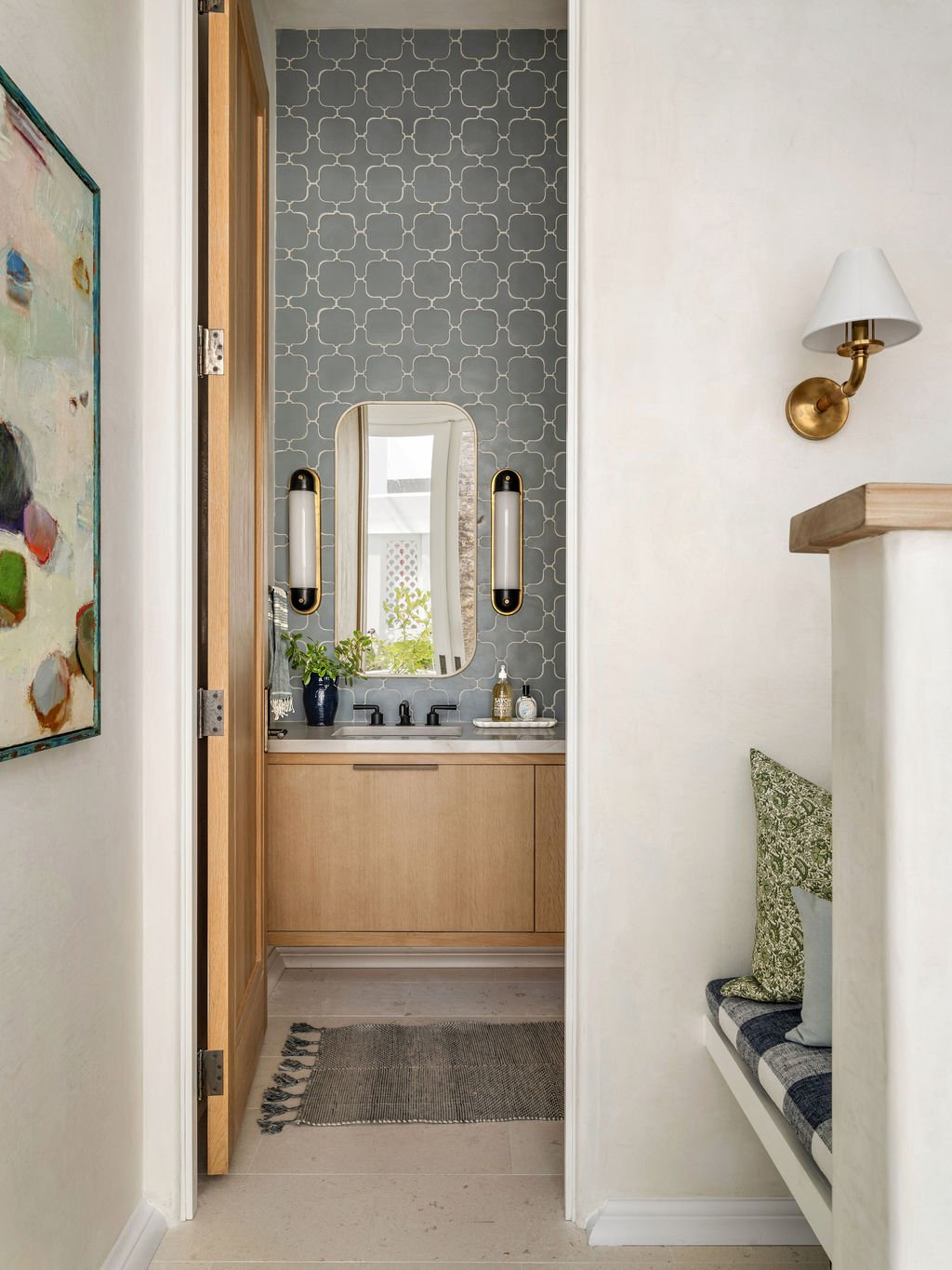 A small bathroom with a vanity, mirror, and wall-mounted lights, visible through an open door from a hallway with a built-in seating area and wall sconce.
