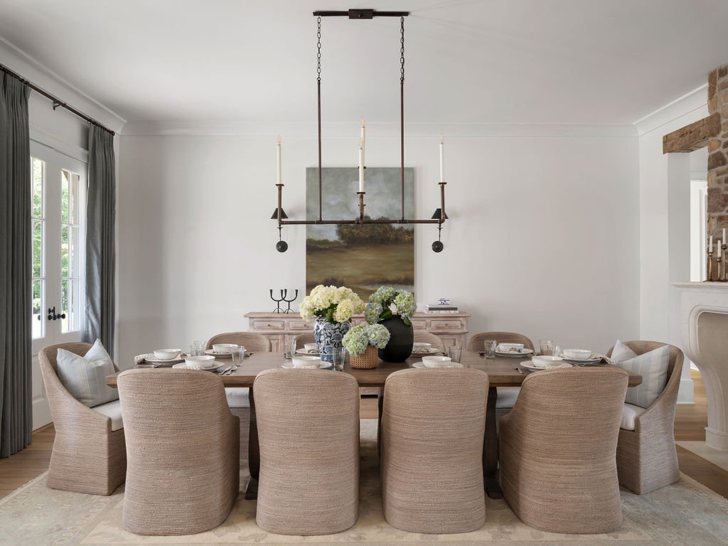 A dining room with a wooden table, beige upholstered chairs, white tableware, and floral centerpieces. The room has white walls, a large window with gray curtains, a stone fireplace, and a chandelier with candles hanging above the table.