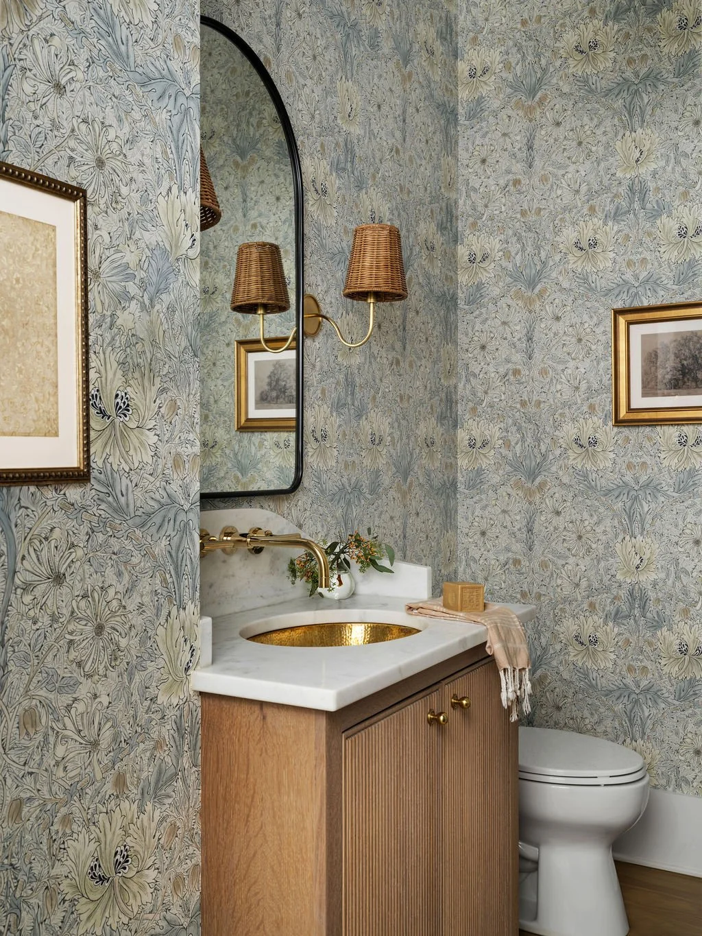 A bathroom with floral wallpaper, a mirror, a gold-colored sink, and a small cabinet with two doors. There are framed pictures on the wall, and a wall-mounted light fixture with two woven shades. A small vase with flowers, a soap dish, and a towel ar