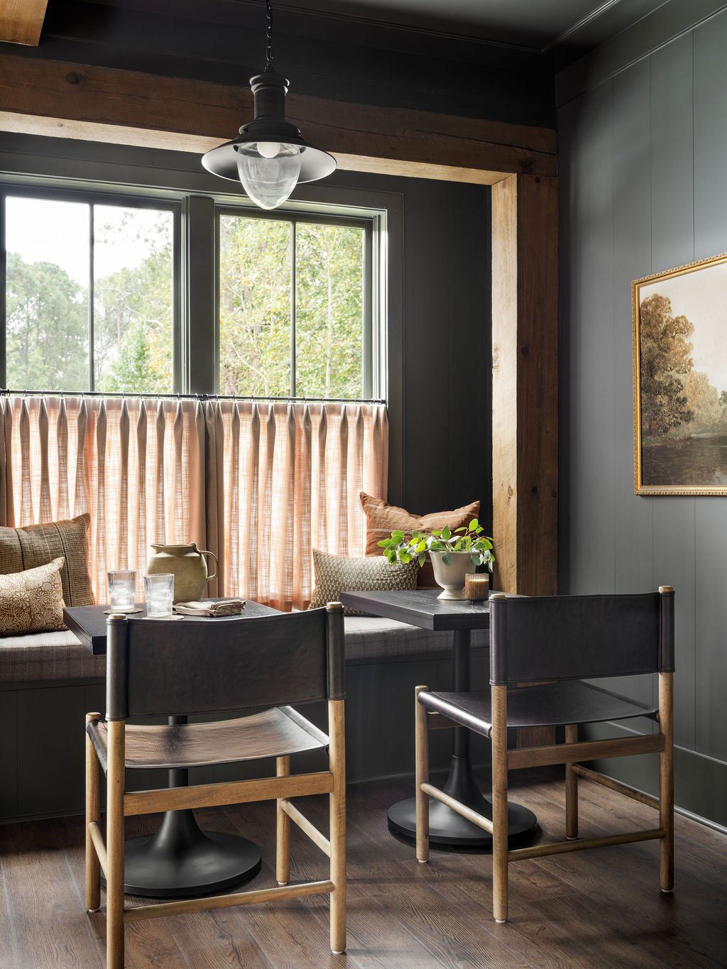 A cozy dining nook with a large window adorned with pink curtains, a built-in bench with pillows, a dark wooden table, black chairs with wooden legs, decorative items including a plant, pottery, and glasses, and landscape artwork on the wall.