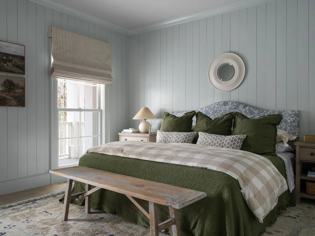 A bedroom with blue walls, a window with a beige Roman shade, a bed with green, gray, and blue bedding, a white bedside table with a lamp, and a wooden bench at the foot of the bed.