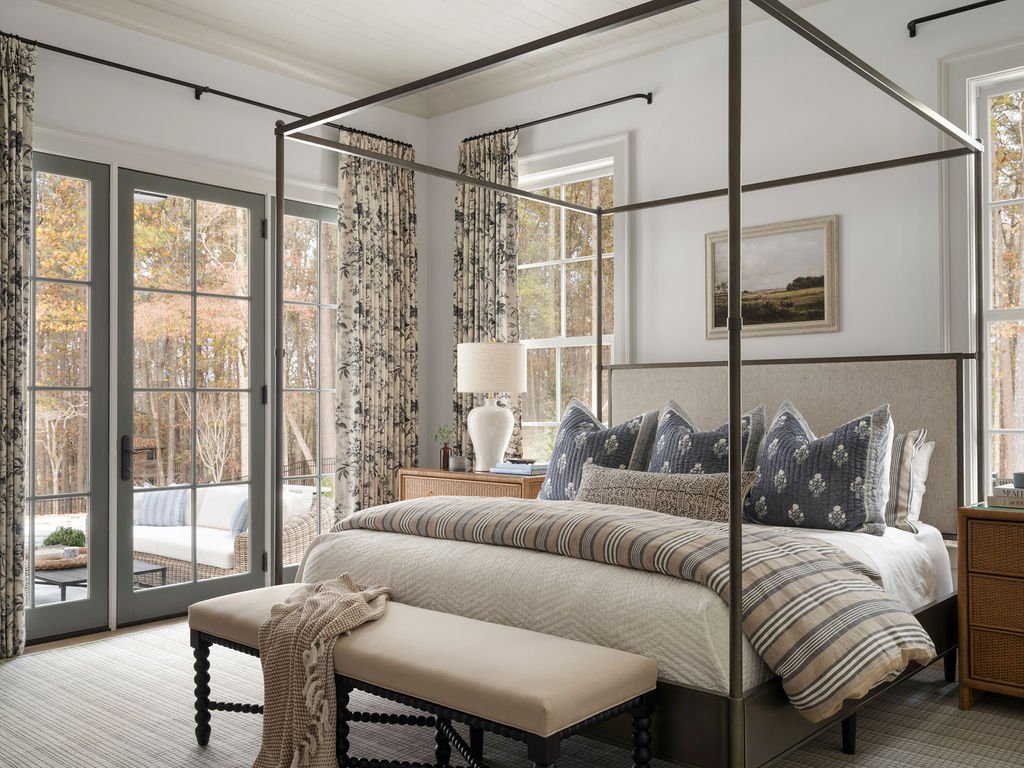 Bedroom with large glass doors, a canopy bed, and a sitting bench at the foot of the bed. Outside patio with seating visible through the doors.