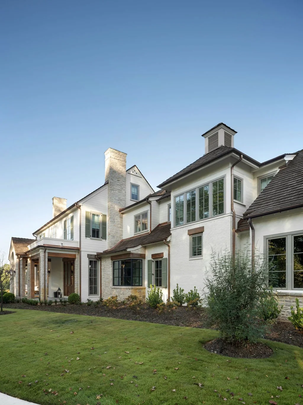 A large, modern house with white brick exterior, multiple large windows, a sloped roof, and landscaping including grass and small shrubs under a clear blue sky.