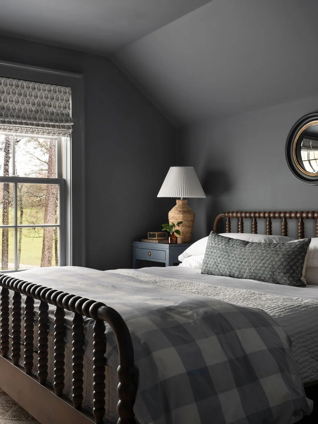 A cozy bedroom with a large window showing trees outside, a bed with a checkered blanket and decorative pillow, a nightstand with a lamp, a clock, and a small plant.