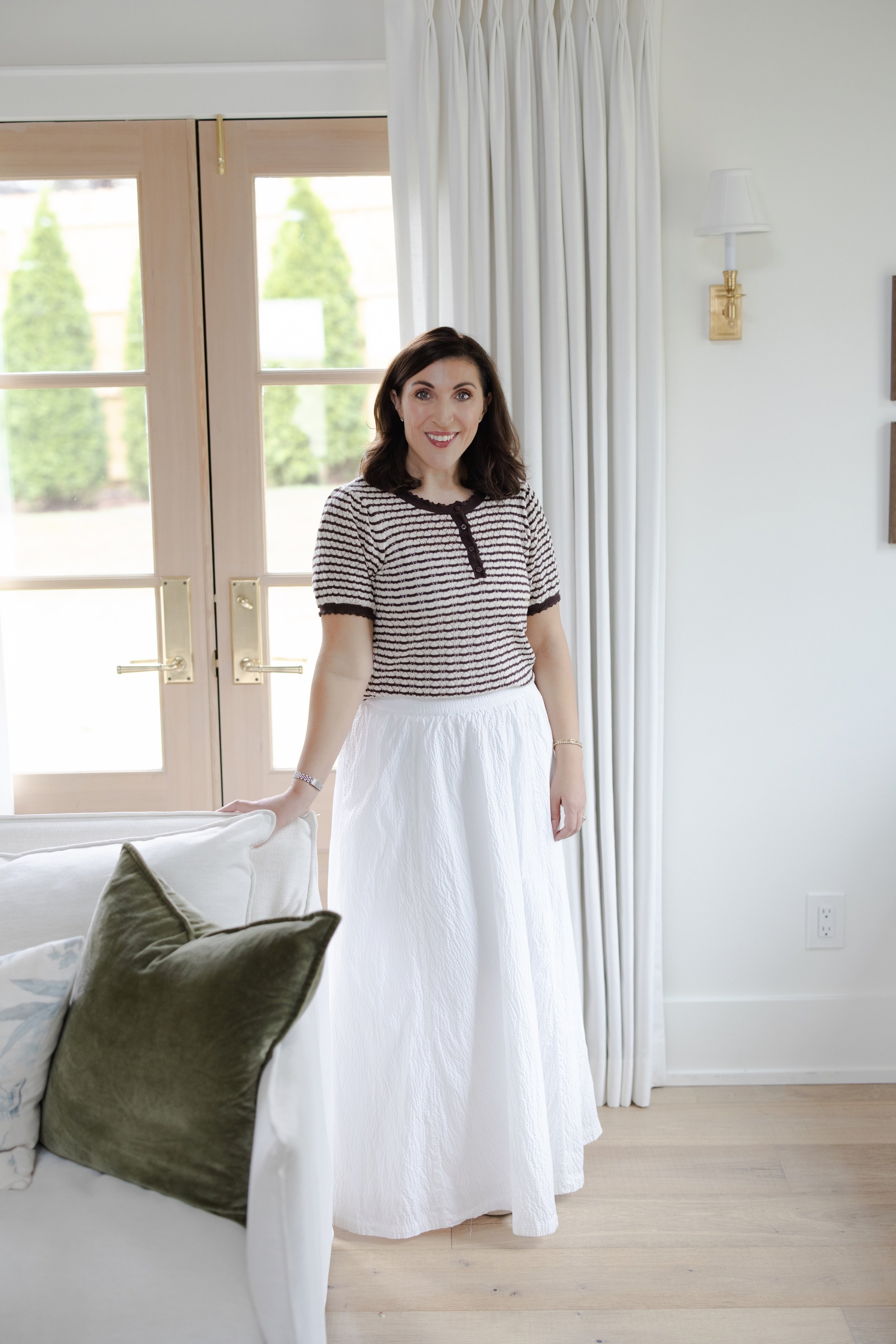 A woman with dark brown hair, wearing a black and white striped top and a long white skirt, standing indoors next to a sofa with green and patterned pillows, in front of glass doors with white curtains.