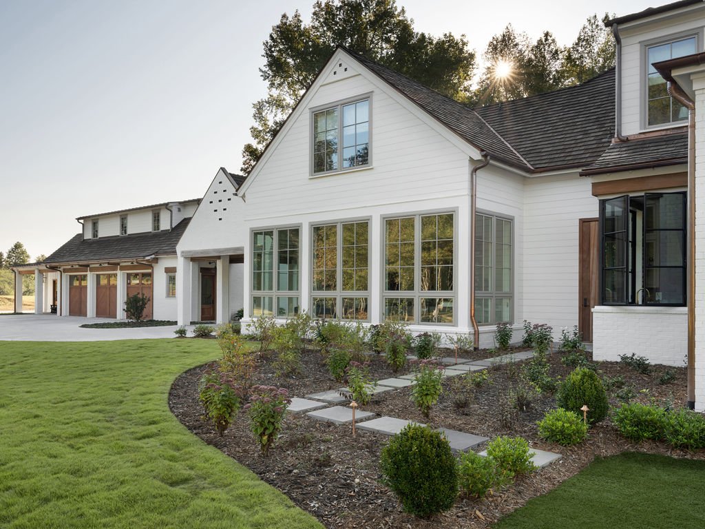 A modern white house with large windows and a sloped roof, surrounded by a landscaped yard with shrubs, flowers, and a grassy lawn, with sunlight peeking through trees in the background.