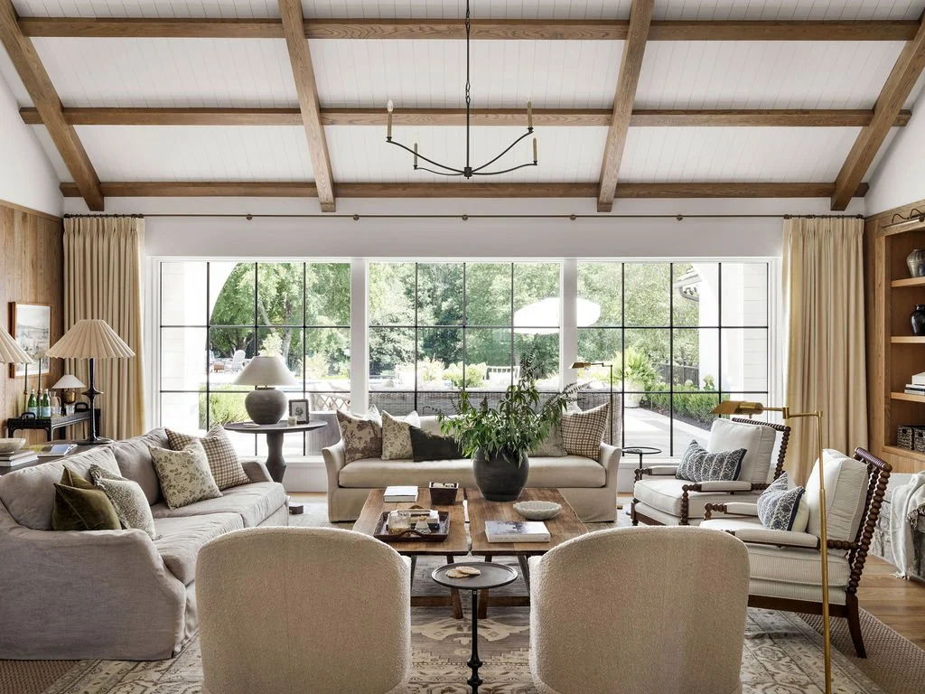 A living room with a vaulted ceiling with wooden beams, large window with curtains, beige sofa and chairs, wooden coffee table, and decorative pillows. There are lamps and plants, with a view of greenery outside.