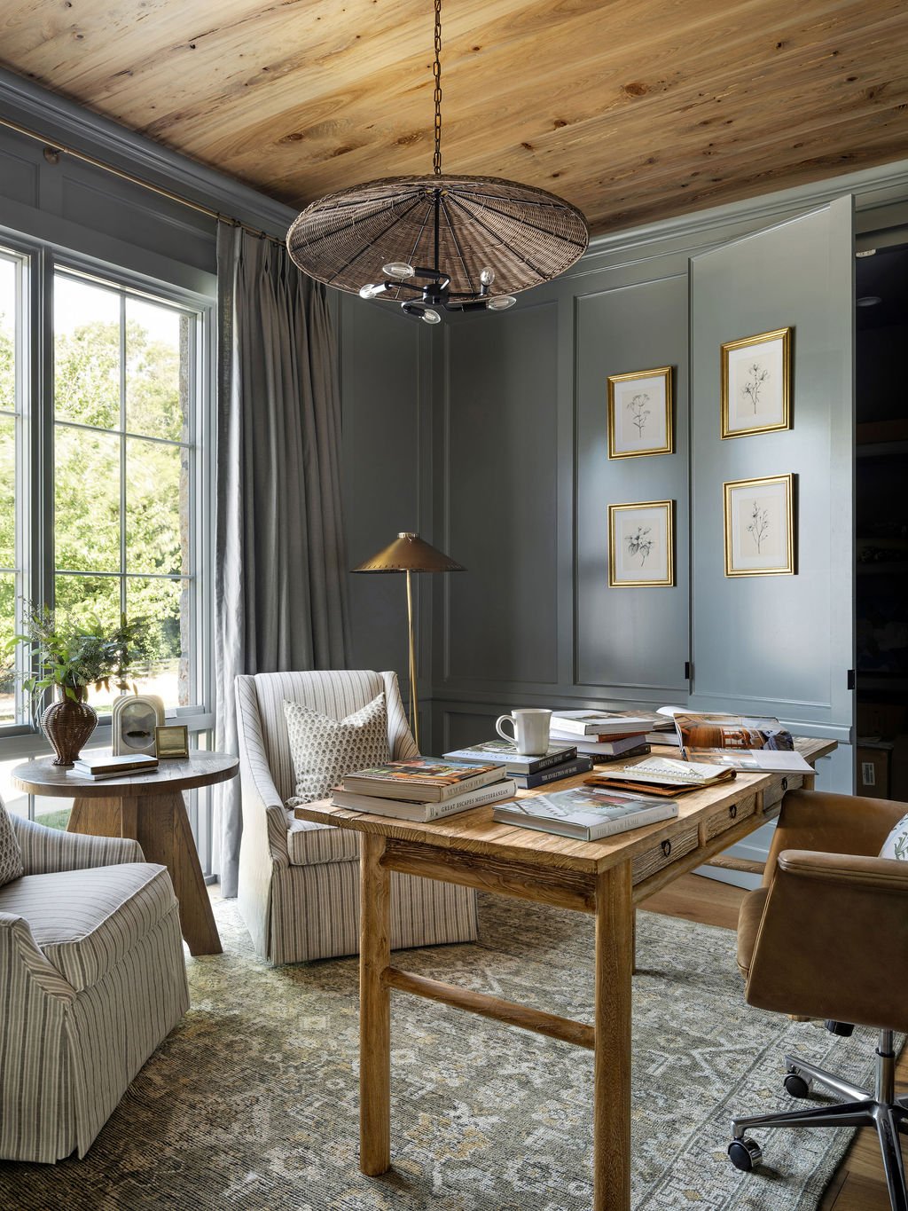 A cozy home office with a wood ceiling, large window with curtains, and a desk cluttered with books, a mug, and papers. There are striped armchairs, a side table with a plant and picture frame, a floor lamp, and framed botanical art on the wall.