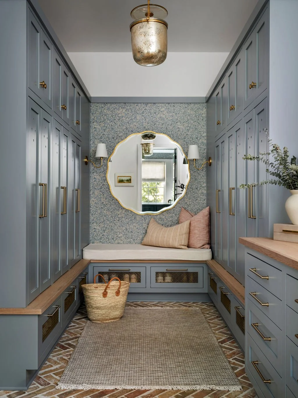 A cozy mudroom with blue cabinetry, a built-in bench with pillows, a large mirror, and a basket on the floor. The room has patterned wallpaper, wall sconces, and a central pendant light.