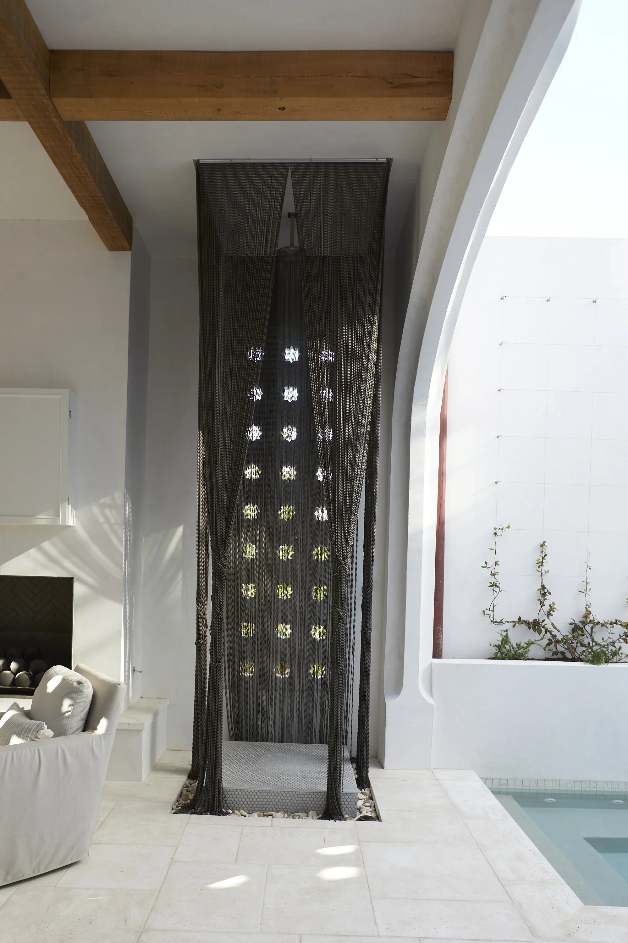Outdoor shower with black curtain beside a white wall and pool, with a sitting area nearby.