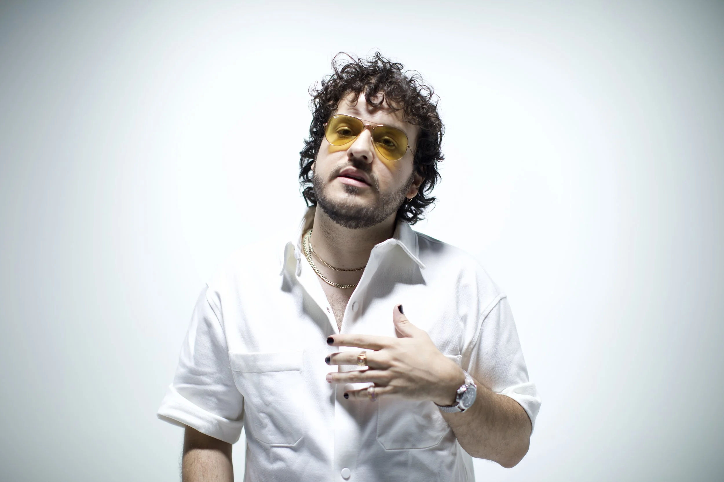 Stephan Stanzione shown with curly hair and a beard wearing yellow-tinted sunglasses, a white button-up shirt, multiple necklaces, rings, and a watch, standing against a plain white background.