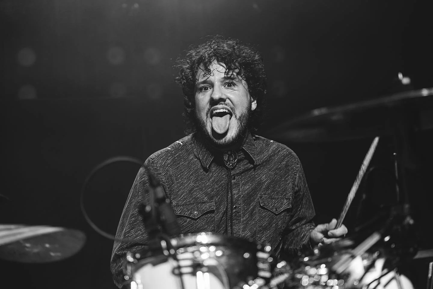 Black and white photo of Stephan Stanzione shown with curly hair and a beard sticking out his tongue, holding a drumstick, with drums in the foreground.