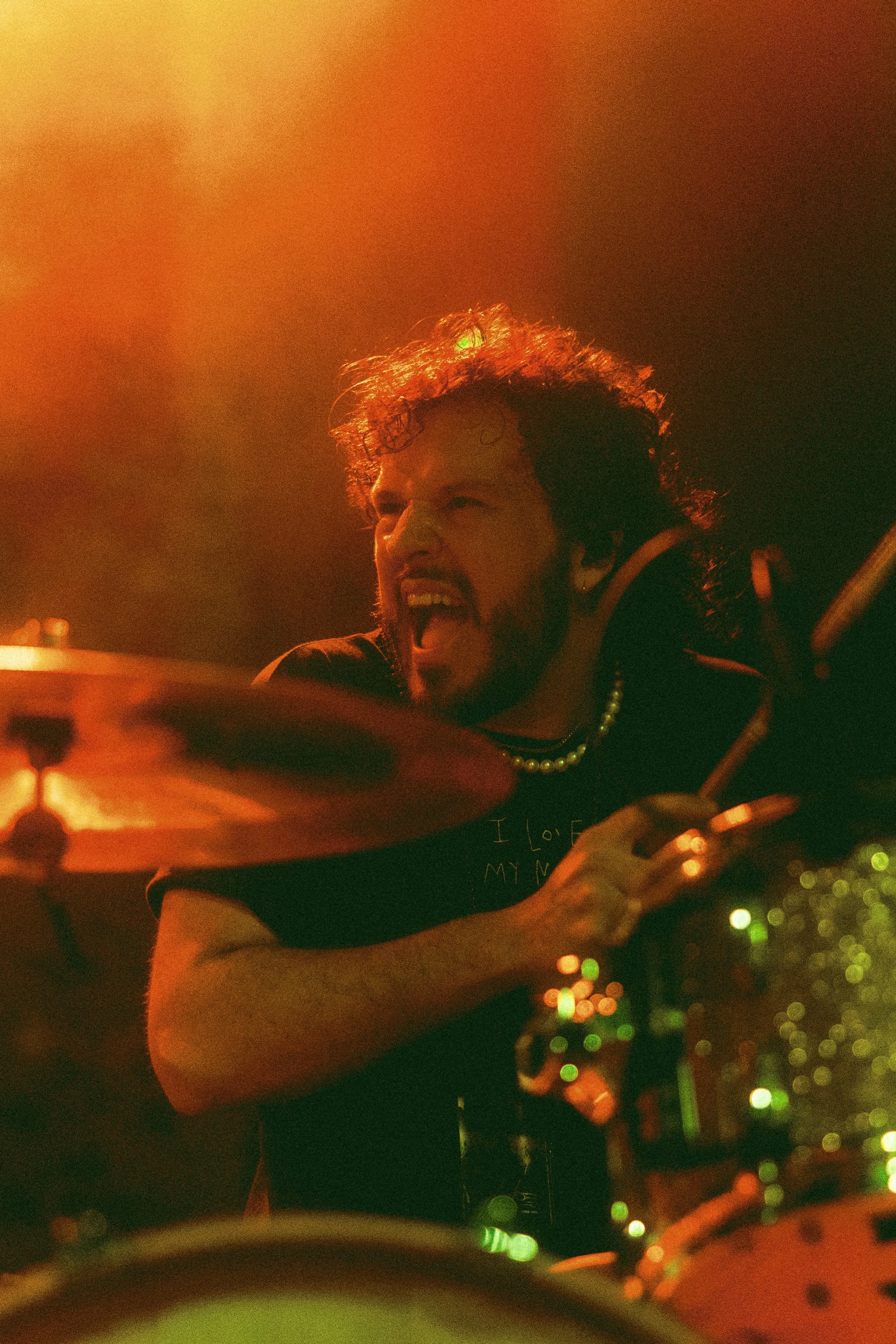 Stephan Stanzione passionately playing drums in a dimly lit setting with red and orange lighting.