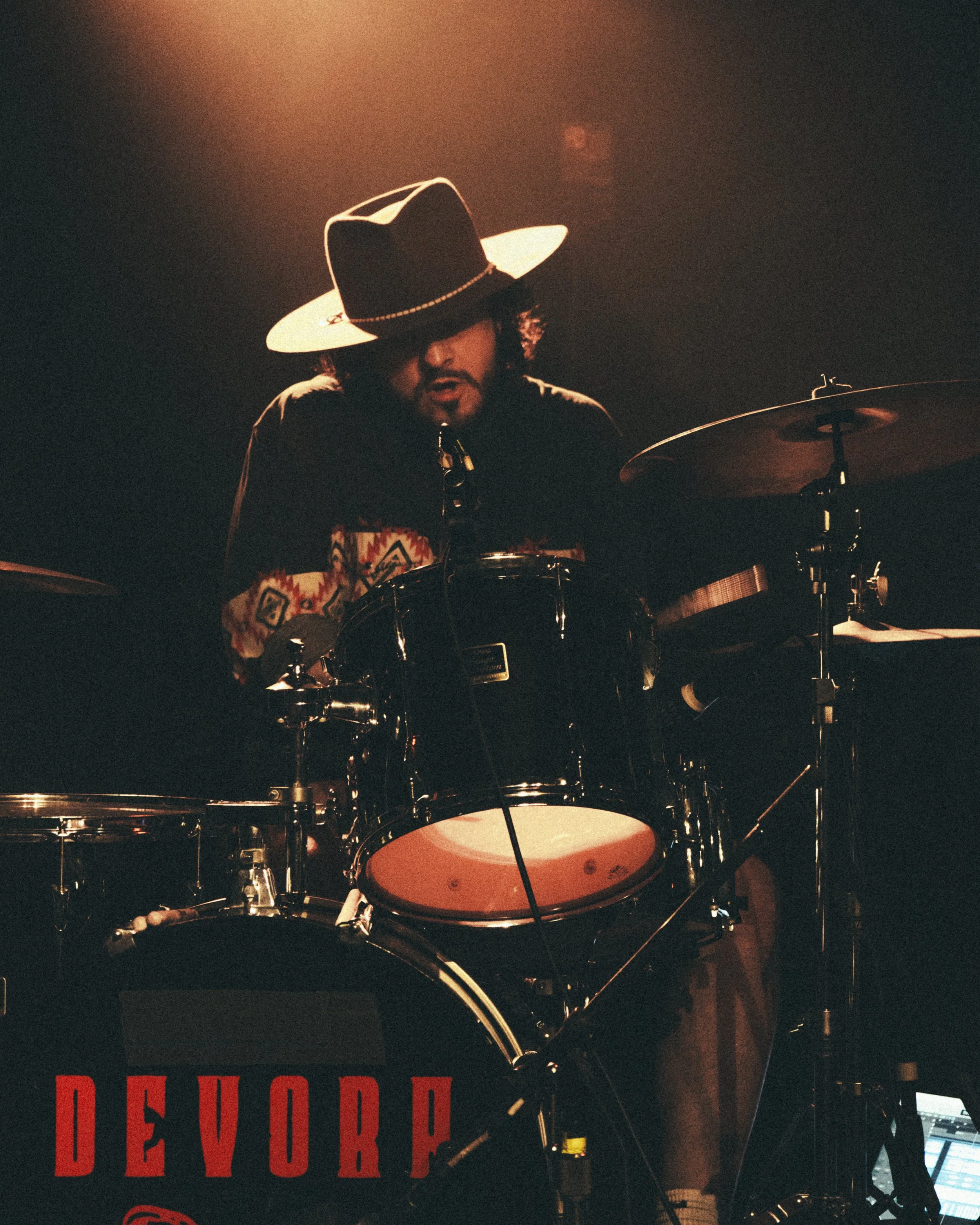 Stephan Stanzione wearing a wide-brimmed hat playing a drum set on stage with dim lighting.