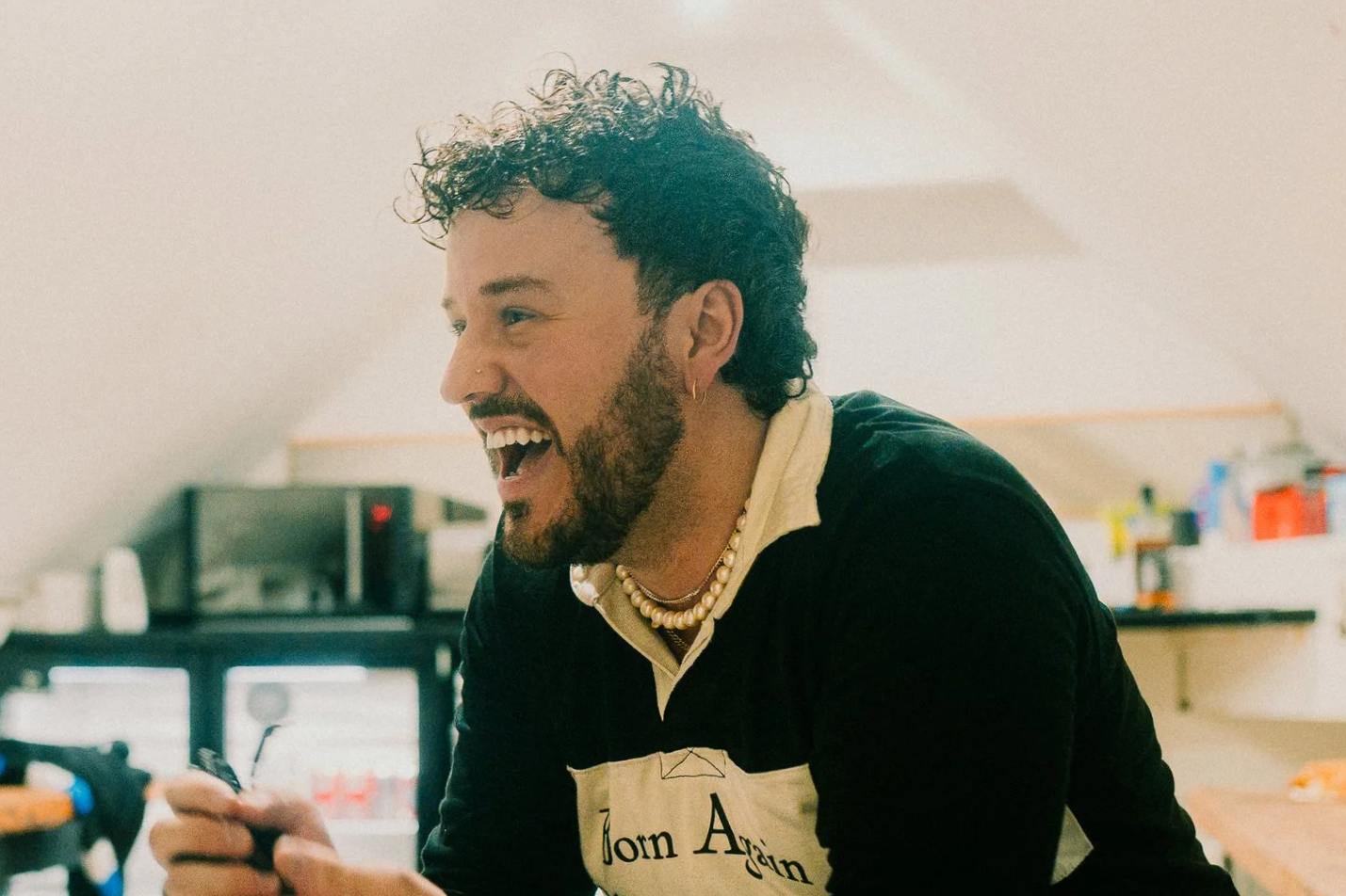 A young man named Stephan stanzione with curly hair, a beard, and earrings, smiling while looking at his phone in a kitchen. This was taken on the Music Of One Tree Hill Tour in Europe with Tyler Hilton and Kate Voegele.