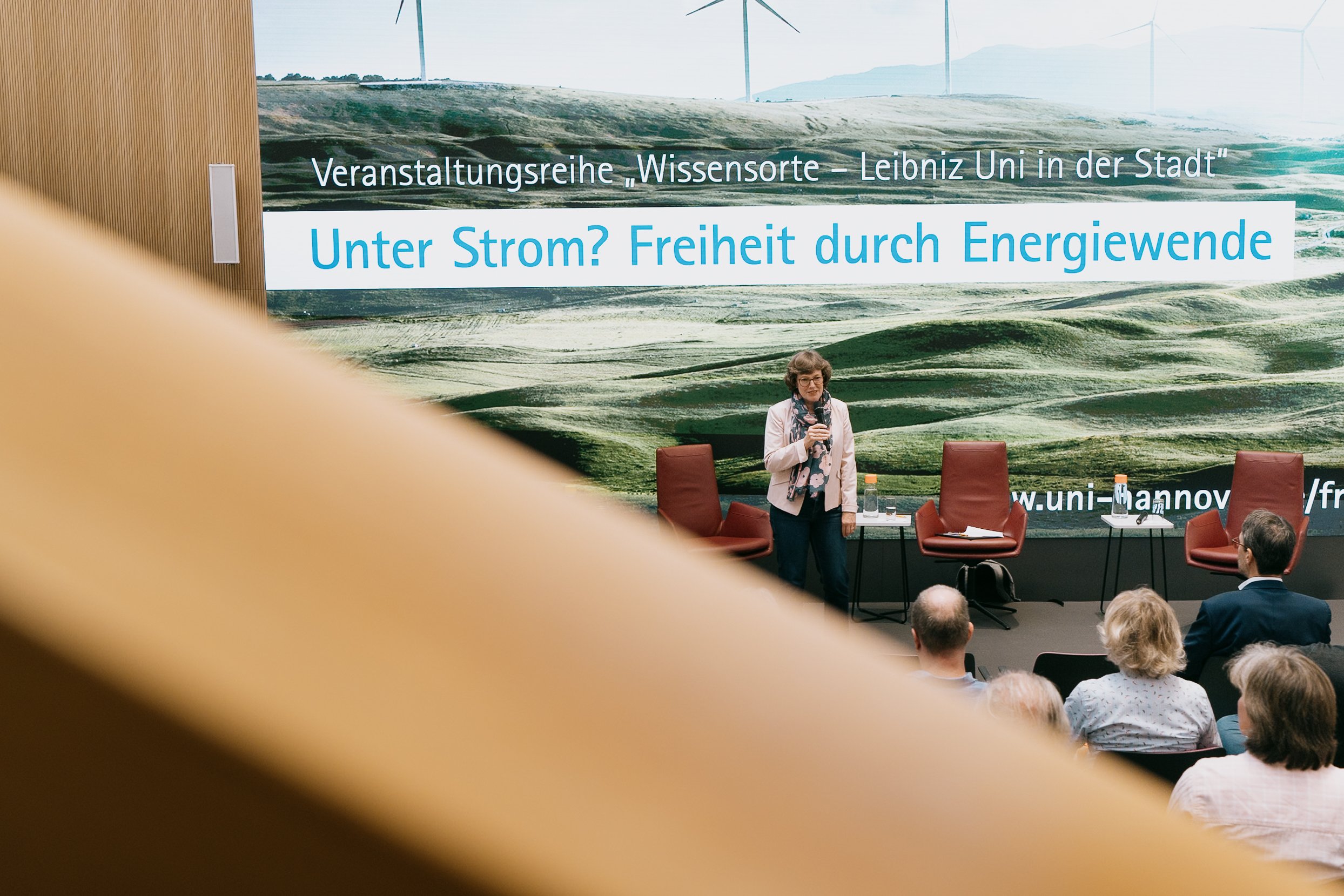 Eine Frau spricht bei einer Veranstaltung über Energiewende vor einem Publikum. Im Hintergrund ist ein großer Bildschirm mit einem Landschaftsfoto und einem Text, der sich um Strom und Energiefreiheit dreht.