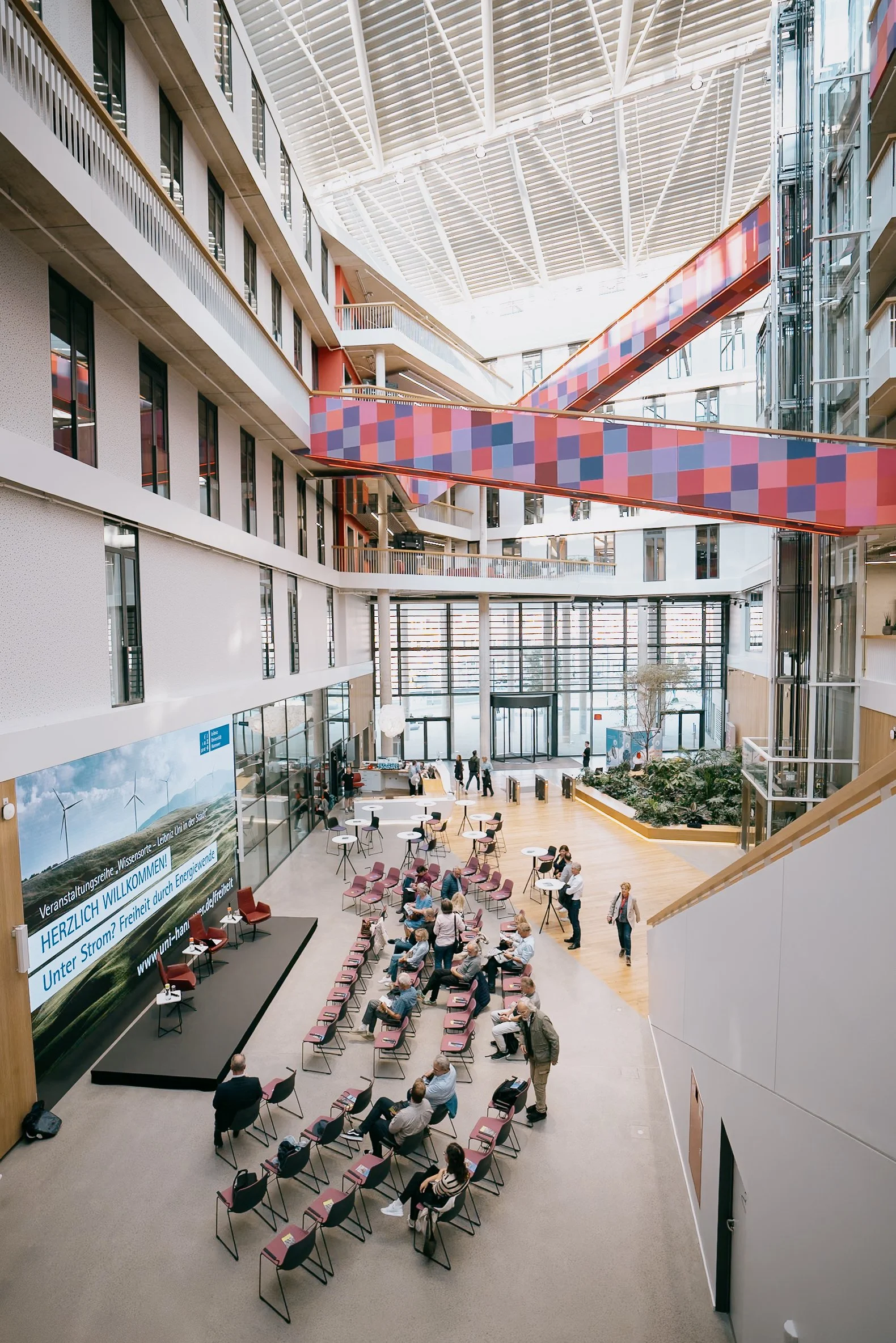 Innenaufnahme eines modernen öffentlichen Gebäudes mit mehreren Stockwerken, einem Vortragsbereich mit Stühlen, einigen Menschen, die sitzen oder stehen, und einer großen LED-Anzeige an der Wand.