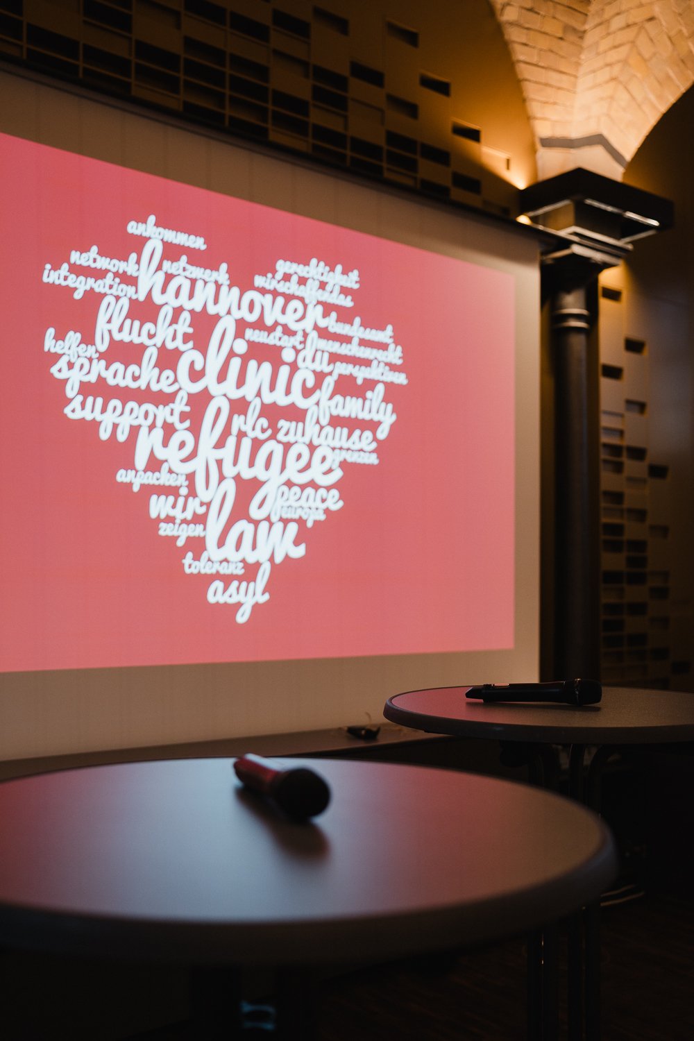 Ein Projektorbild zeigt ein Herz aus deutschen Worten wie 'gemeinsam', 'flucht', 'support', 'peace' und 'tolerance' auf einem pinken Hintergrund in einem dunklen Raum. Im Vordergrund stehen zwei Tische mit Mikrofonen.