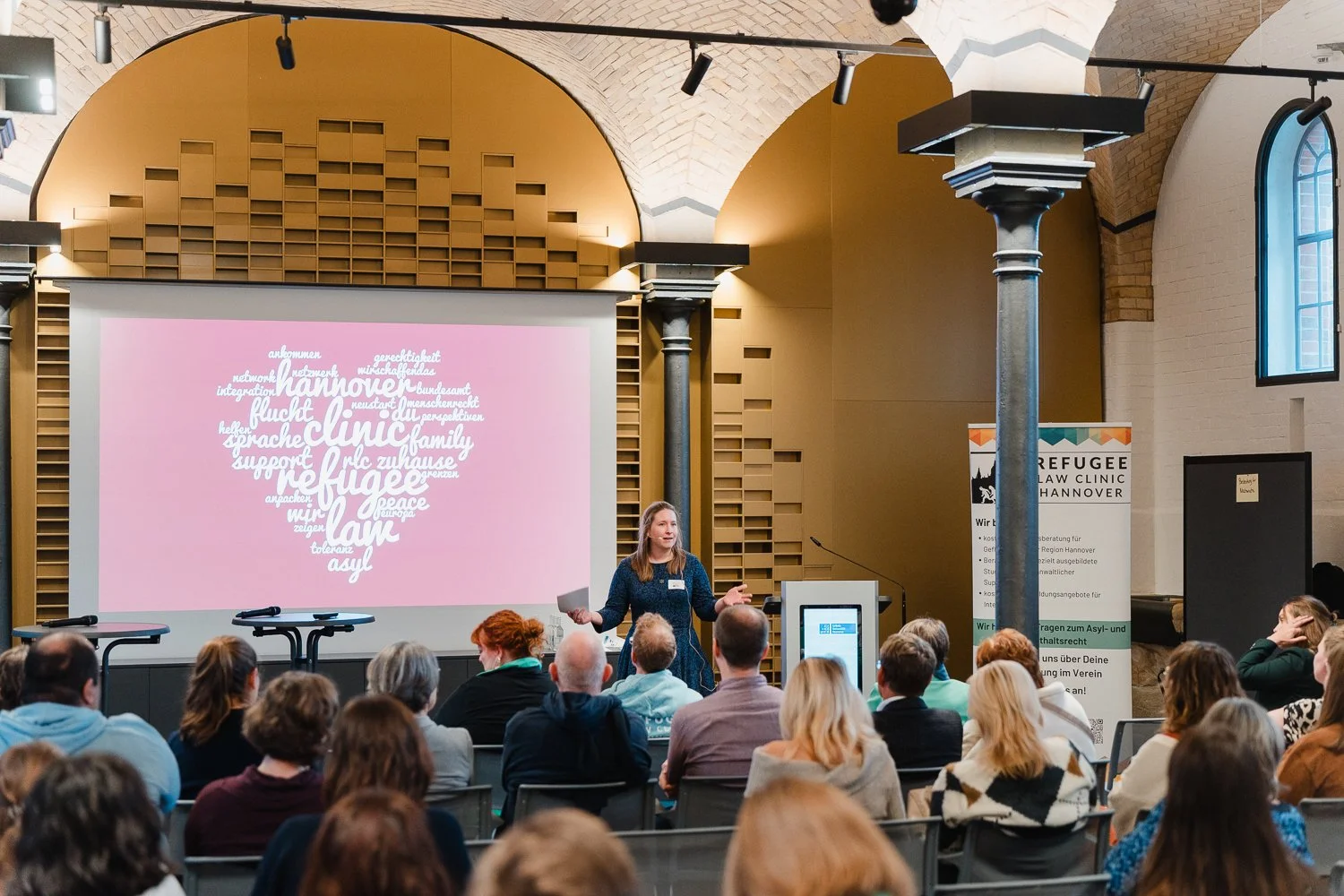 Eine Frau hält einen Vortrag vor einem Publikum in einem großen Raum mit hohen Fenstern und einer großen Projektionsfläche, auf der ein Wortwolkenbild in Herzform zu sehen ist.