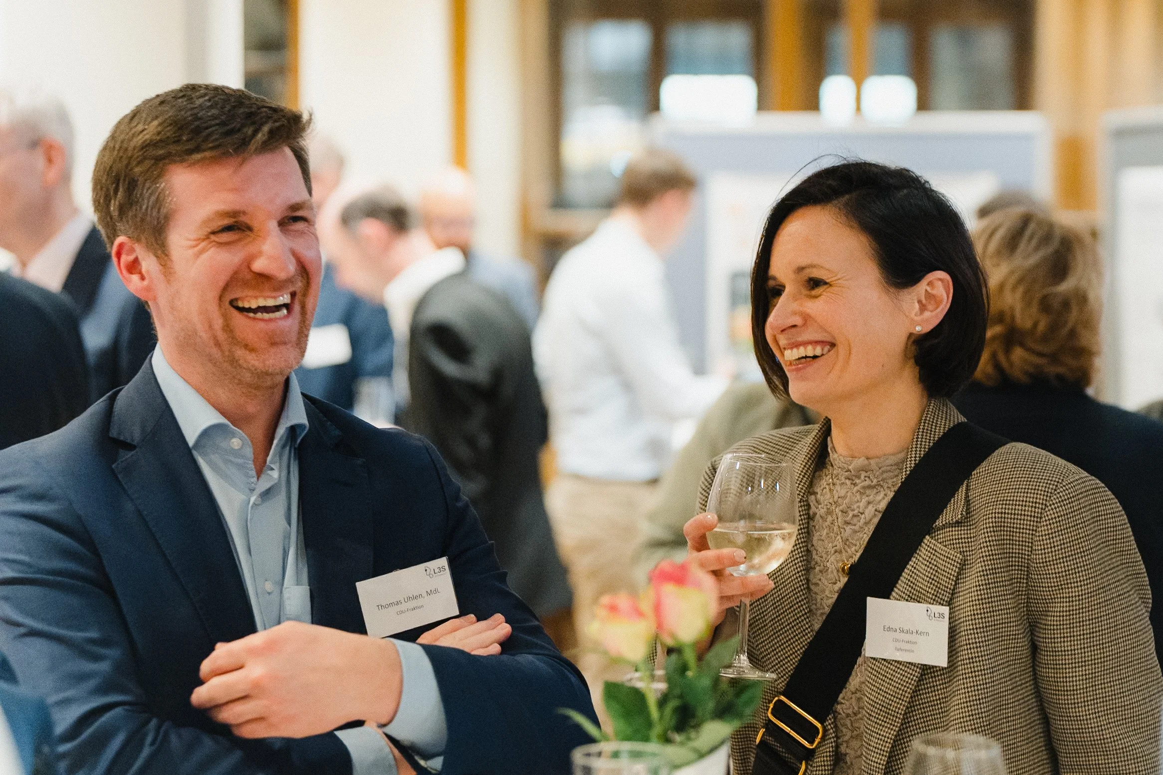 Menschen lachen und unterhalten sich auf einer Netzwerkveranstaltung, mit Namensschildern und Wein in der Hand.