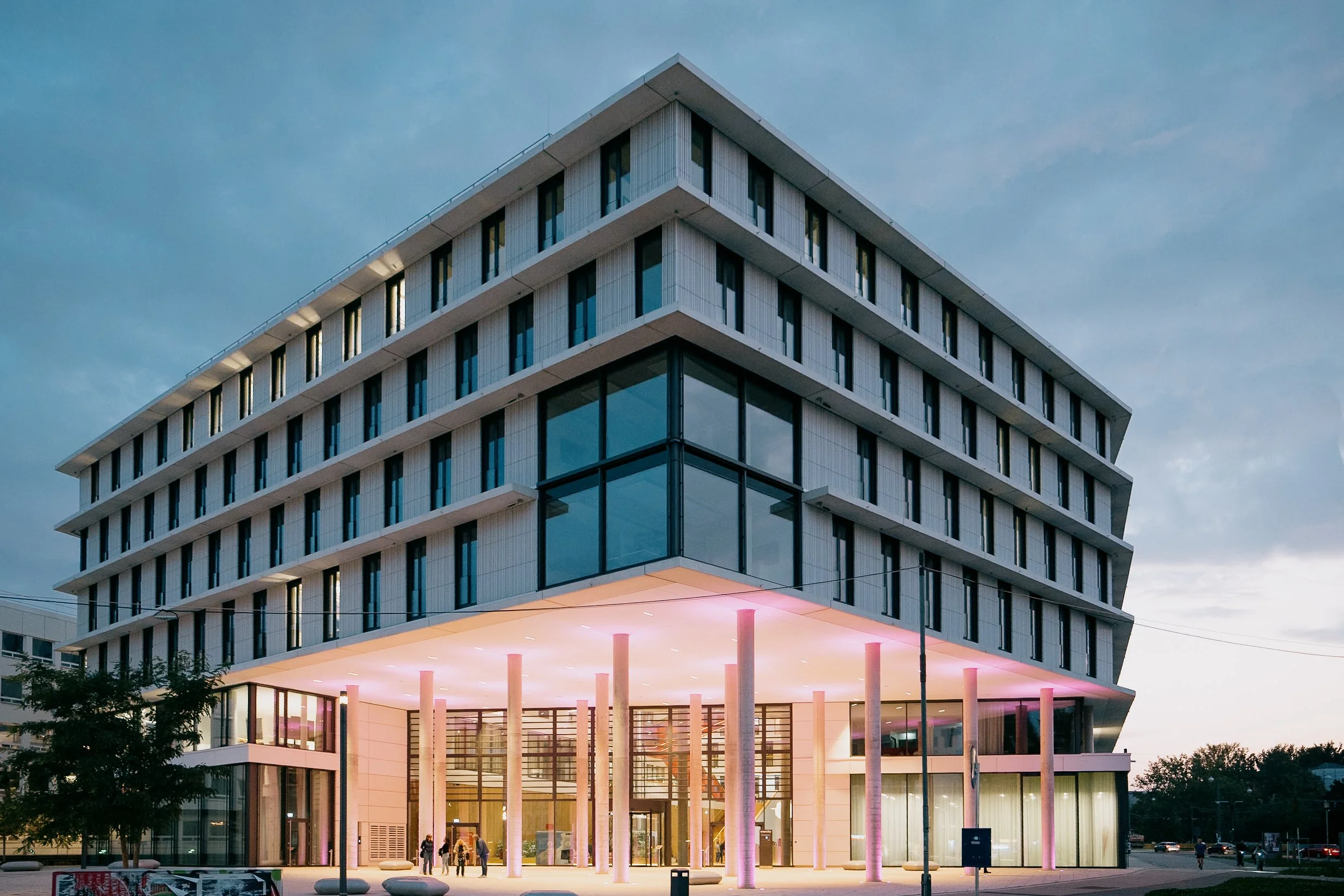 Modernes mehrstöckiges Gebäude mit großen Fenstern und beleuchteter Eingangshalle bei Dämmerung.