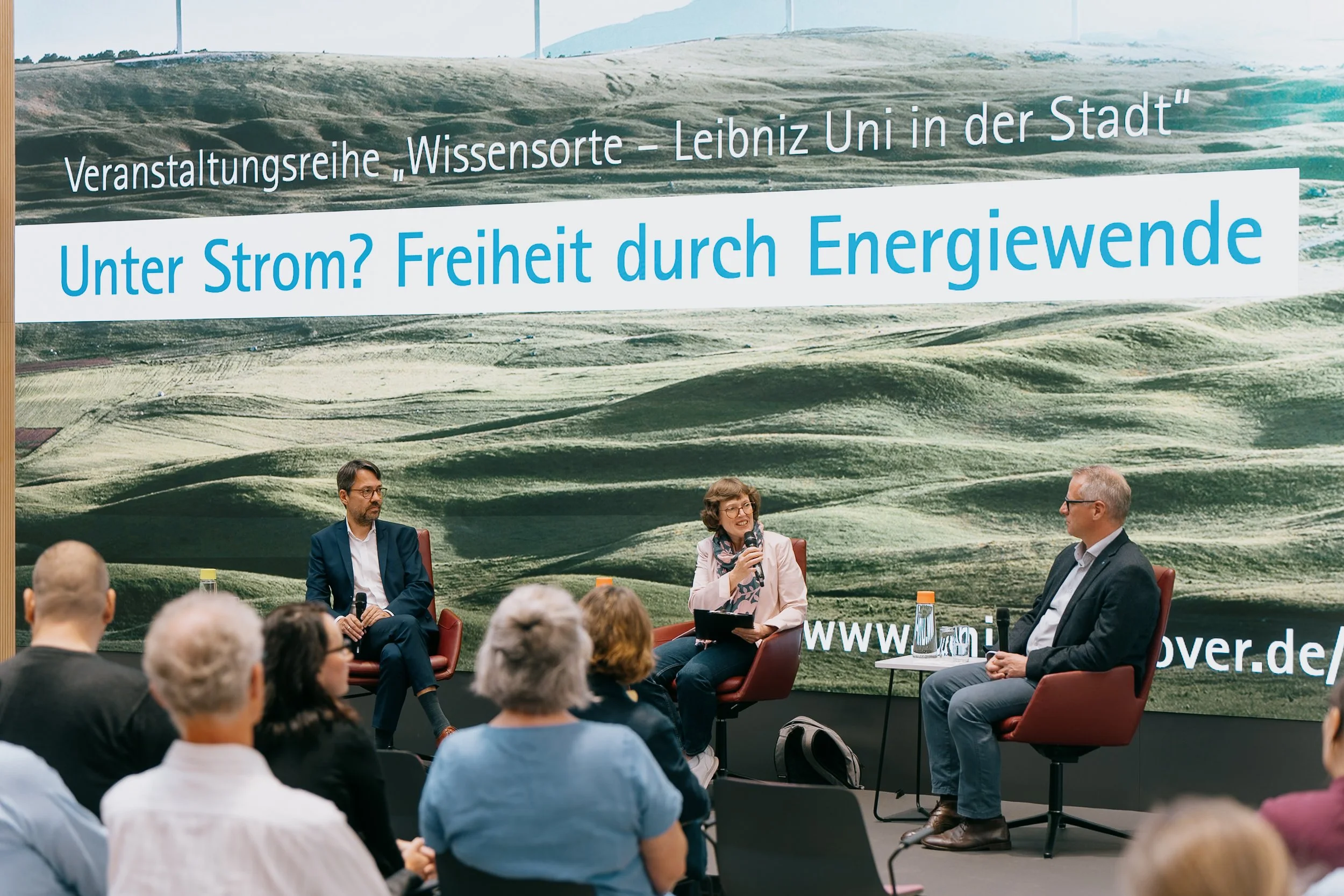 Ein Panel mit drei Personen bei einer Diskussion vor einer großen grünen Landschaft. Das Hintergrundbanner trägt den Titel 'Unter Strom? Freiheit durch Energiewende' und erwähnt eine Veranstaltungsreihe an der Leibniz Universität.