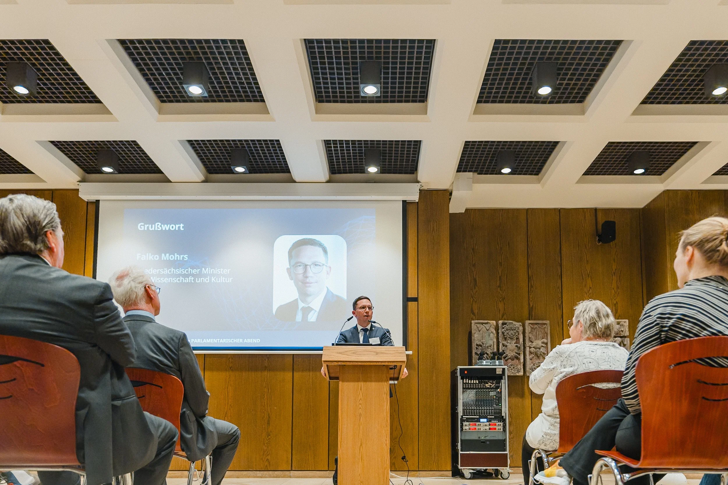 Ein Vortrag in einem Saal mit einem Redner an einem Podium vor einer großen Leinwand. Die Zuschauer sitzen in Reihen und schauen auf die Präsentation, auf der auch sein Bild und Name zu sehen sind.