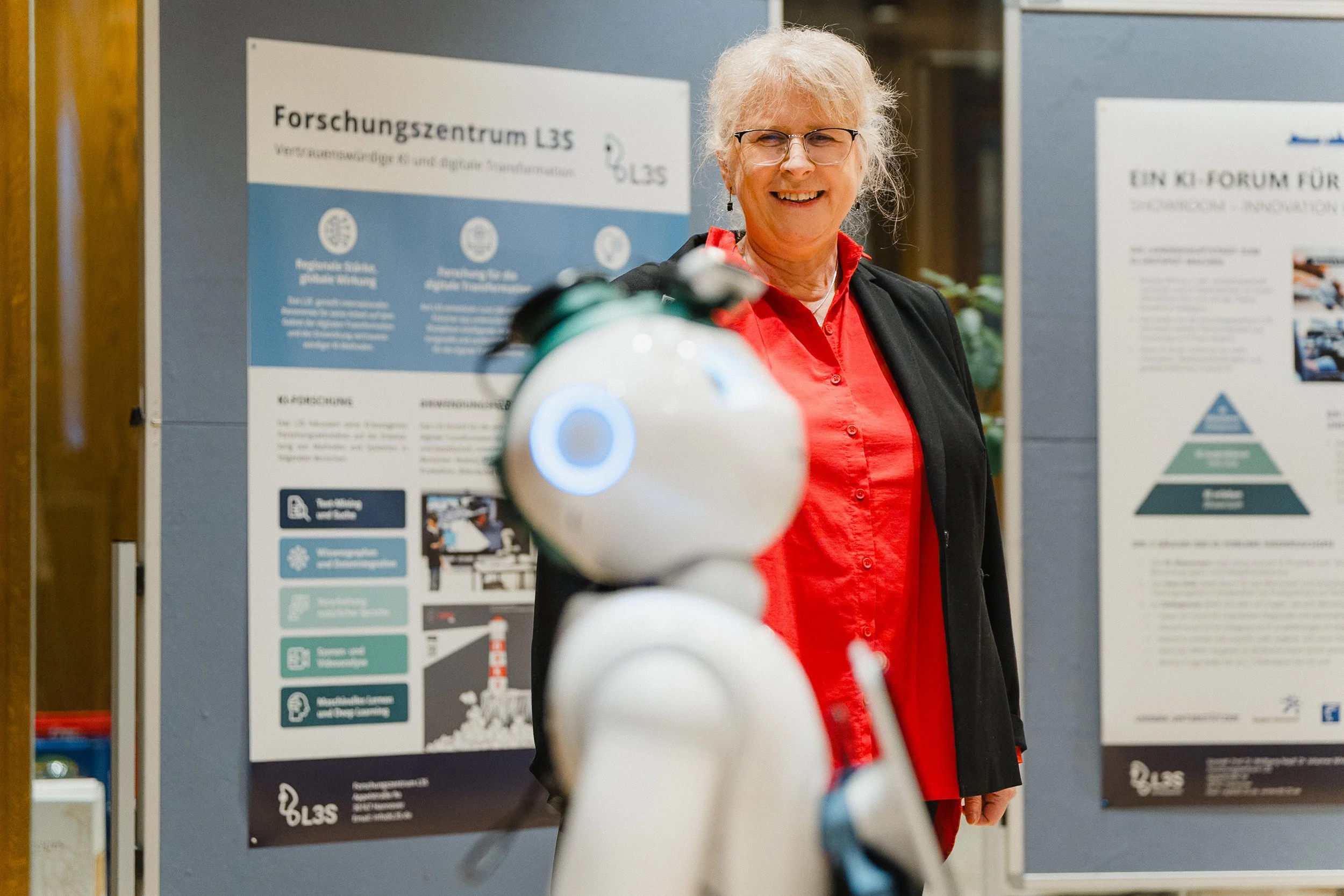 Eine lächelnde ältere Frau mit längeren weißen Haaren, Brille und rotem Hemd steht vor Informationsplakaten, im Vordergrund ist ein humanoider Roboter. Die Szene scheint in einem Forschungszentrum oder einer Konferenz zu sein.