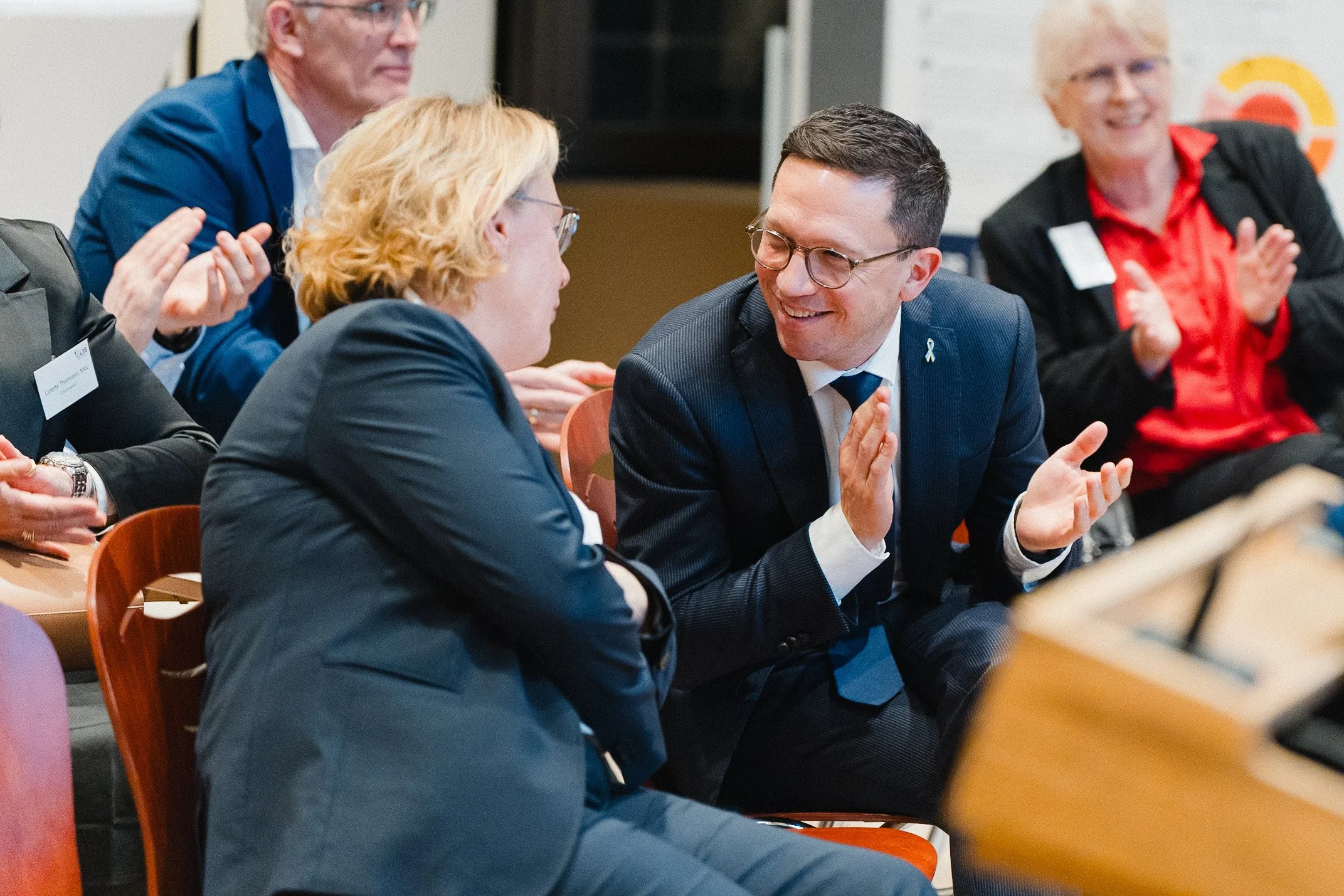 Menschen bei einer Veranstaltung, die sich unterhalten und applaudieren, einige sitzen im Gespräch, einer lächelt und gestikuliert während einer Diskussion.