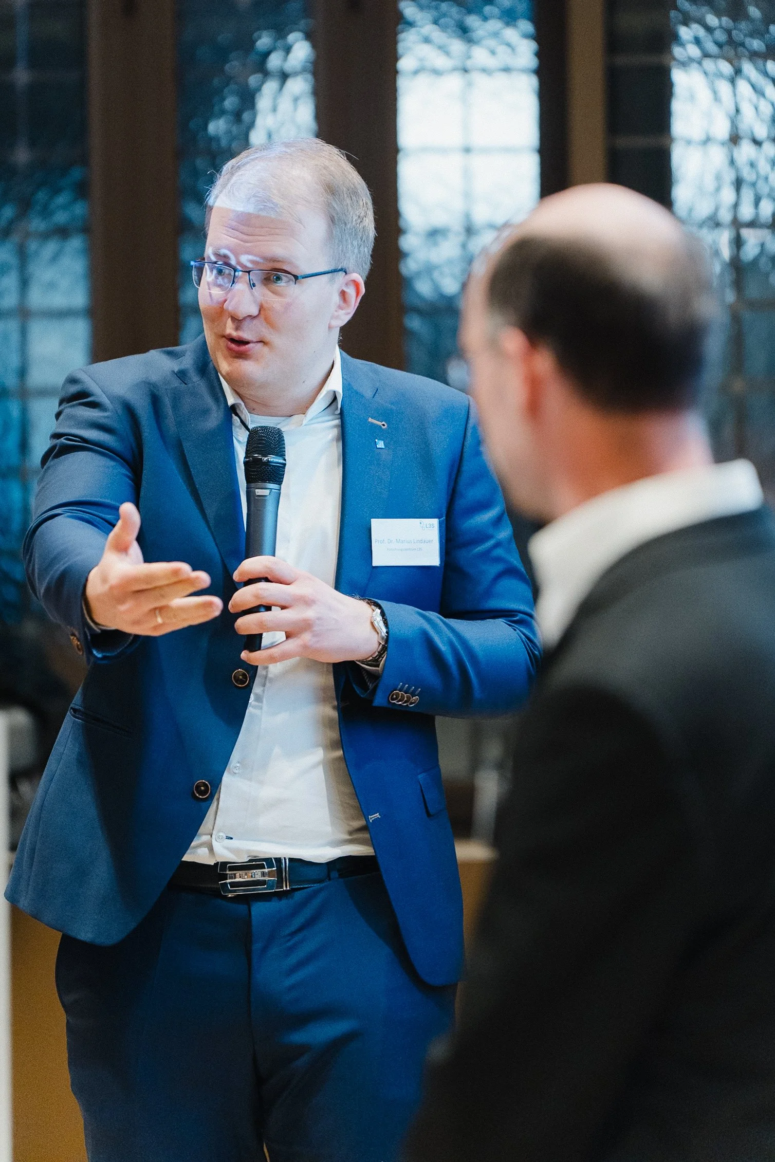 Ein Mann in einem blauen Anzug spricht in einem Raum mit großen Fenstern, während er mit einem Mikrofon spricht.
