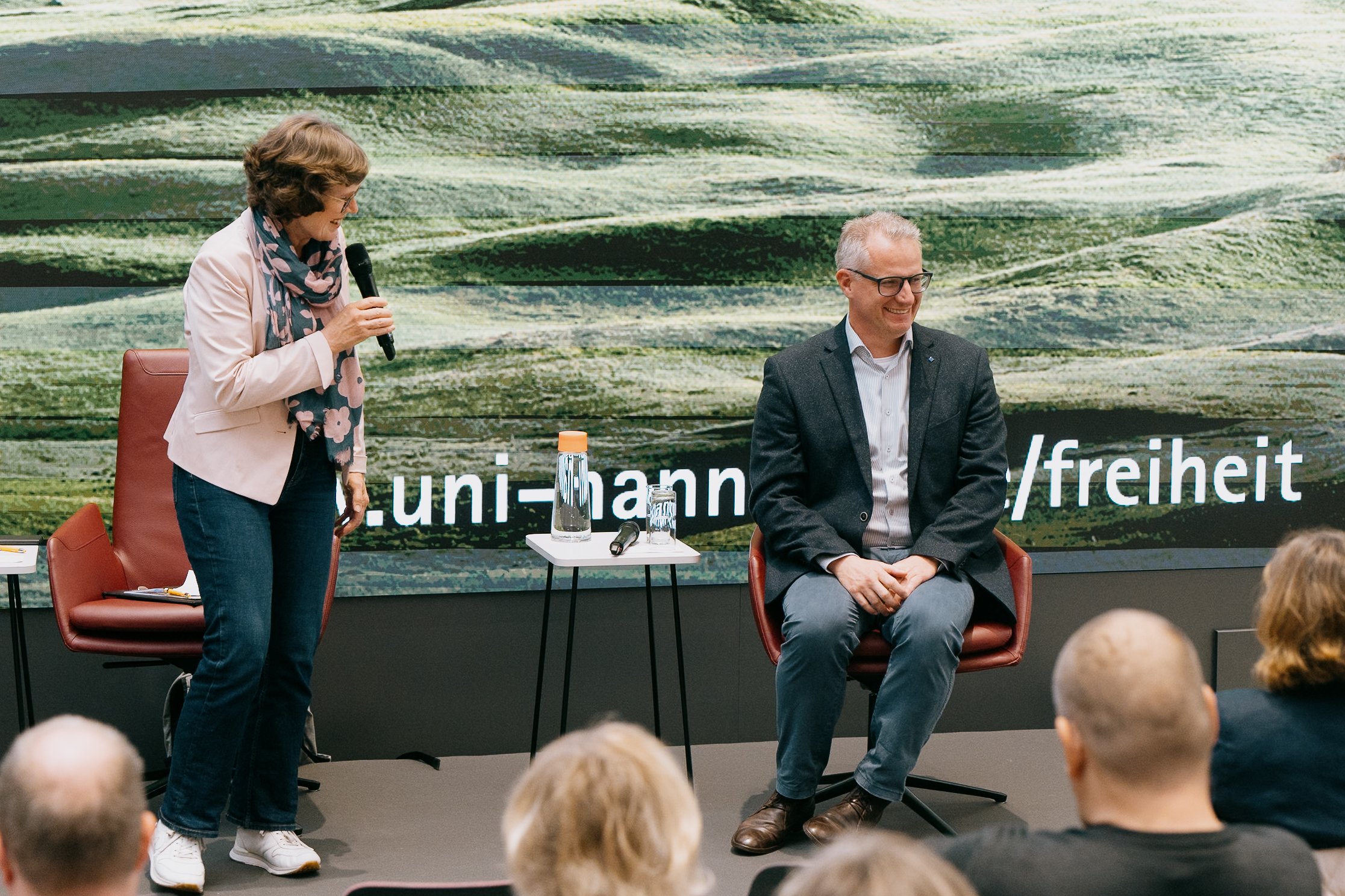 Eine Frau mit einem Mikrofon spricht zu einem Mann, der auf einem Stuhl sitzt; im Hintergrund ein Bildschirm mit Text. Die Szene scheint eine Podiumsdiskussion oder Präsentation zu sein.