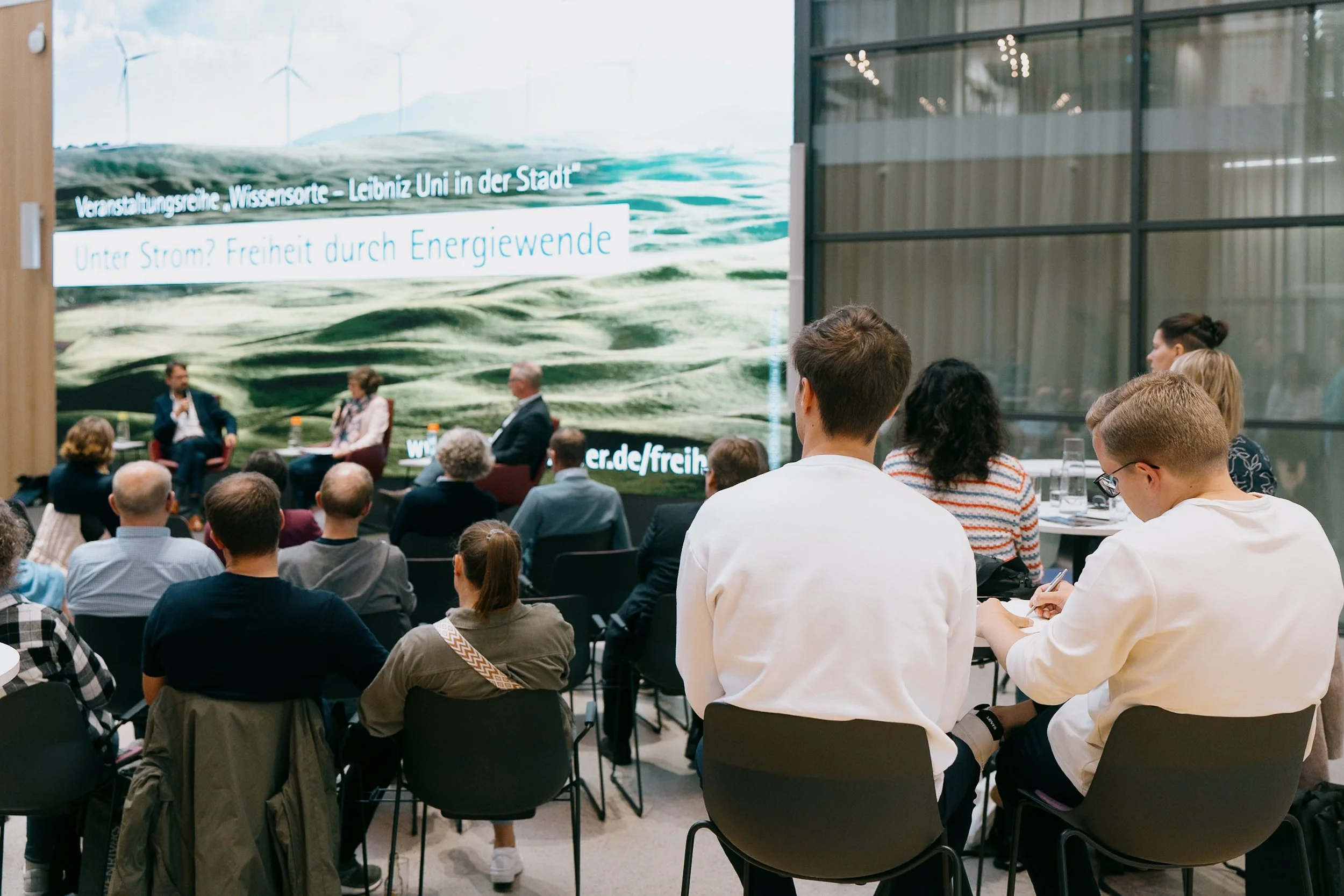 Bürger bei einer Konferenz, die über erneuerbare Energien spricht, vor einem großen Bildschirm mit einem Wasser- und Windpark-Hintergrund.